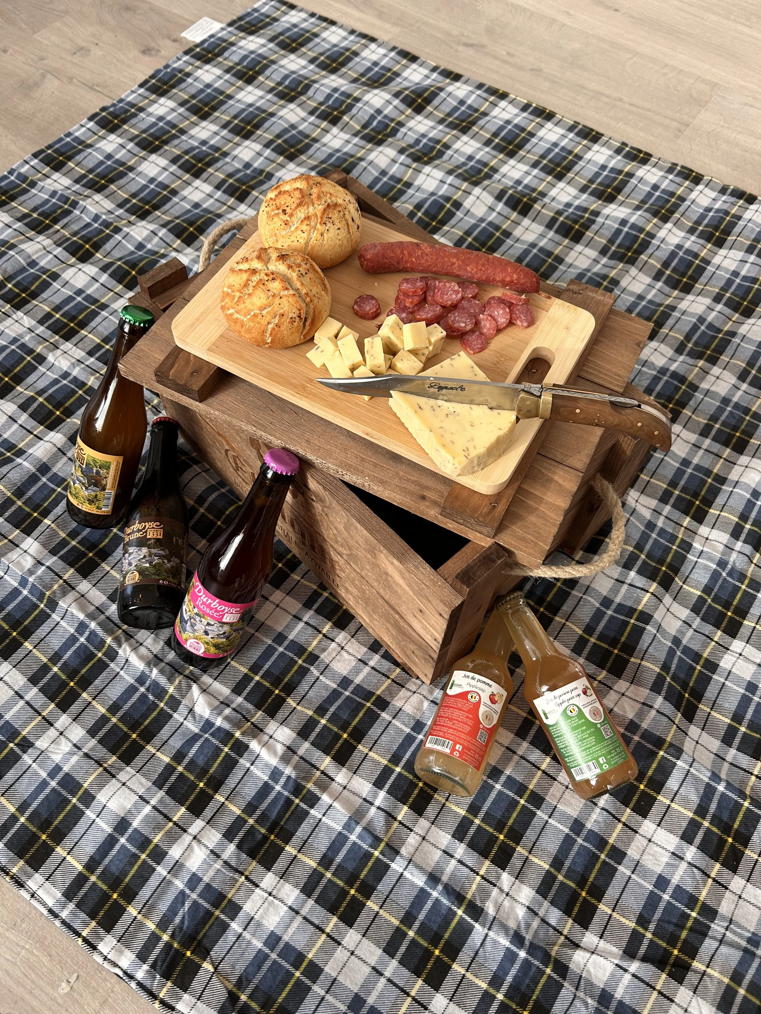 Plate de charcuterie avec pain, fromage, saucisses, et deux bouteilles de boissons fermentées sur une table recouverte d'une nappe à carreaux.