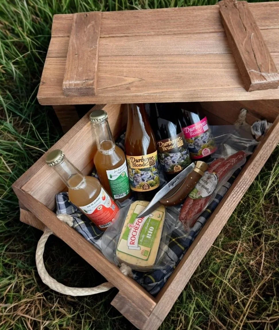 Coffre en bois contenant des bouteilles de bière, un pâté bio, un couteau, et du saucisson, posé dans de l'herbe.