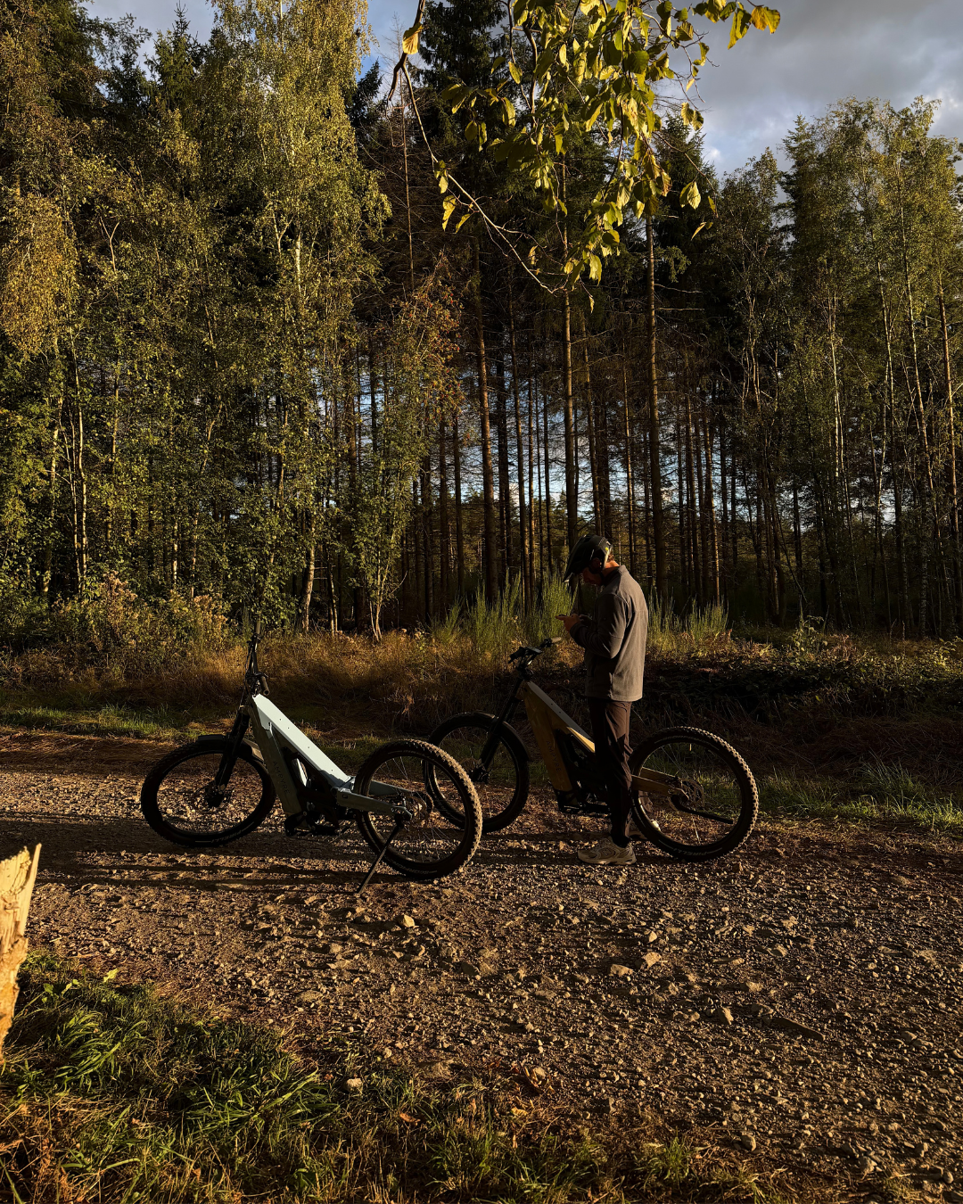 Une personne portant un casque consulte son téléphone, se tenant à côté de deux trottinettes électriques sur un chemin en forêt, avec des arbres verts et un ciel nuageux en arrière-plan.