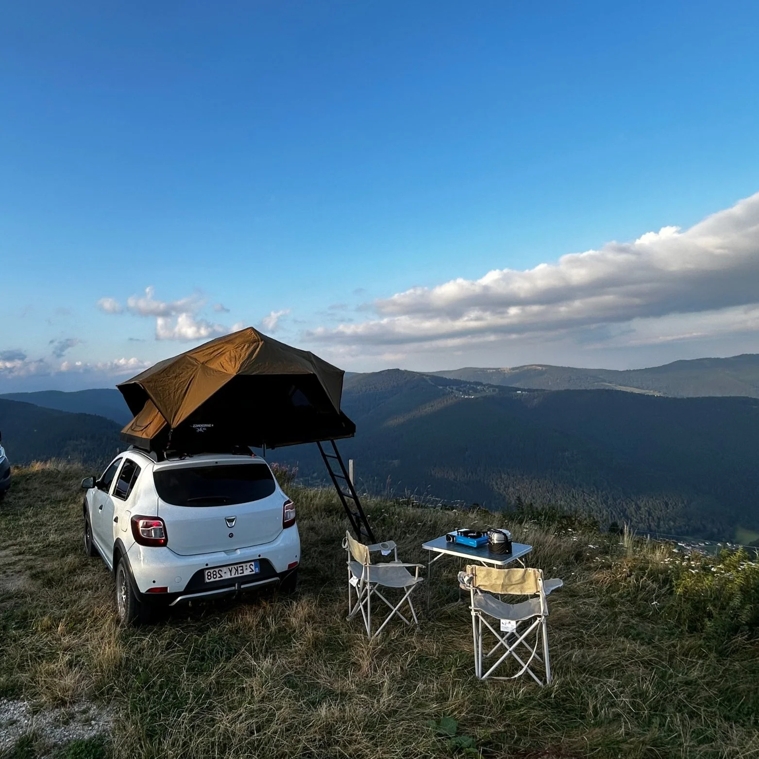 Voiture blanche avec un toit de tente déployé, deux chaises pliantes, une table avec un camping gaz et une marmite, sur une falaise avec vue sur des montagnes et un ciel bleu avec quelques nuages.