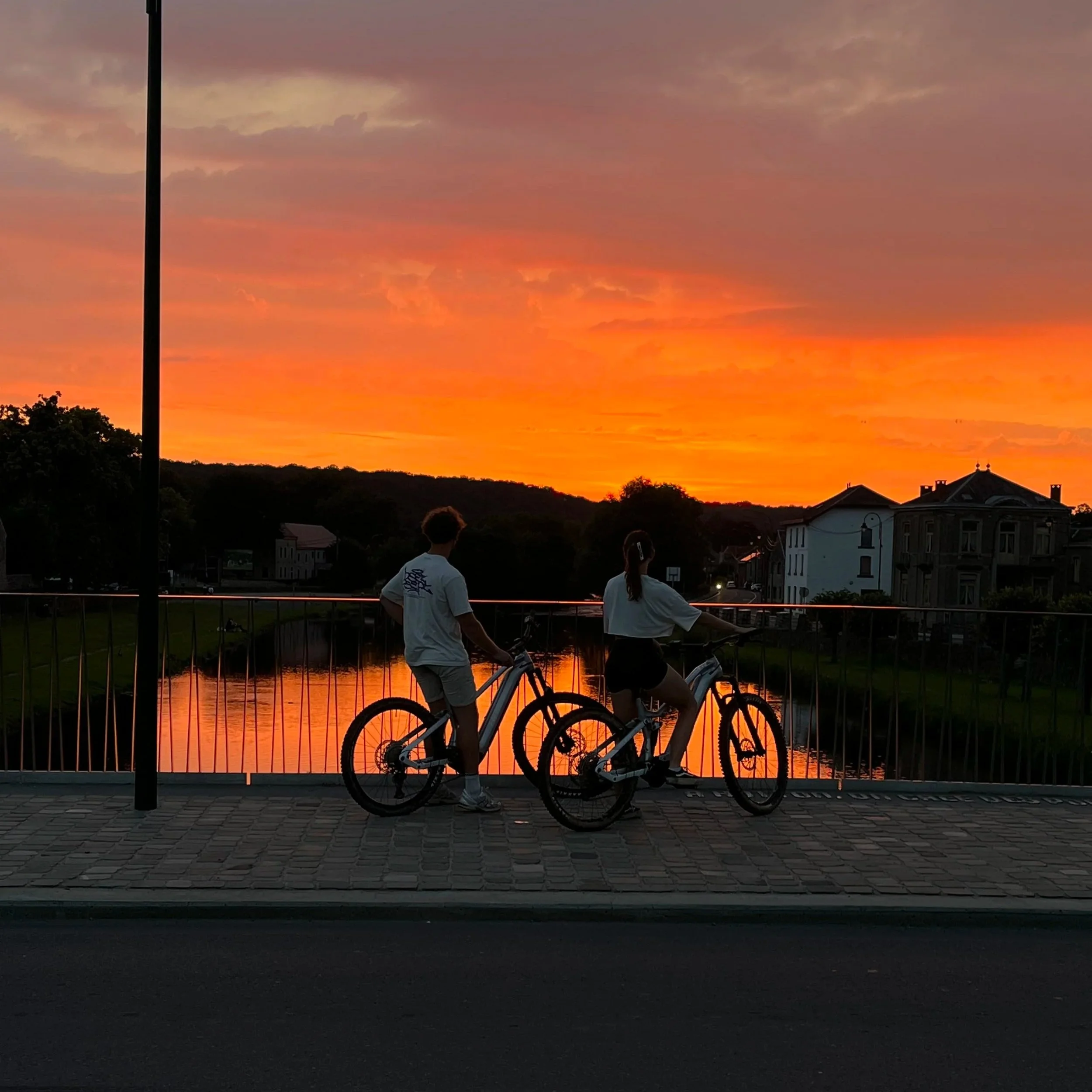 Coucher de soleil orange sur un paysage urbain avec un pont, deux personnes avec des vélos en silhouette, et des bâtiments, vue depuis un trottoir en pavés.