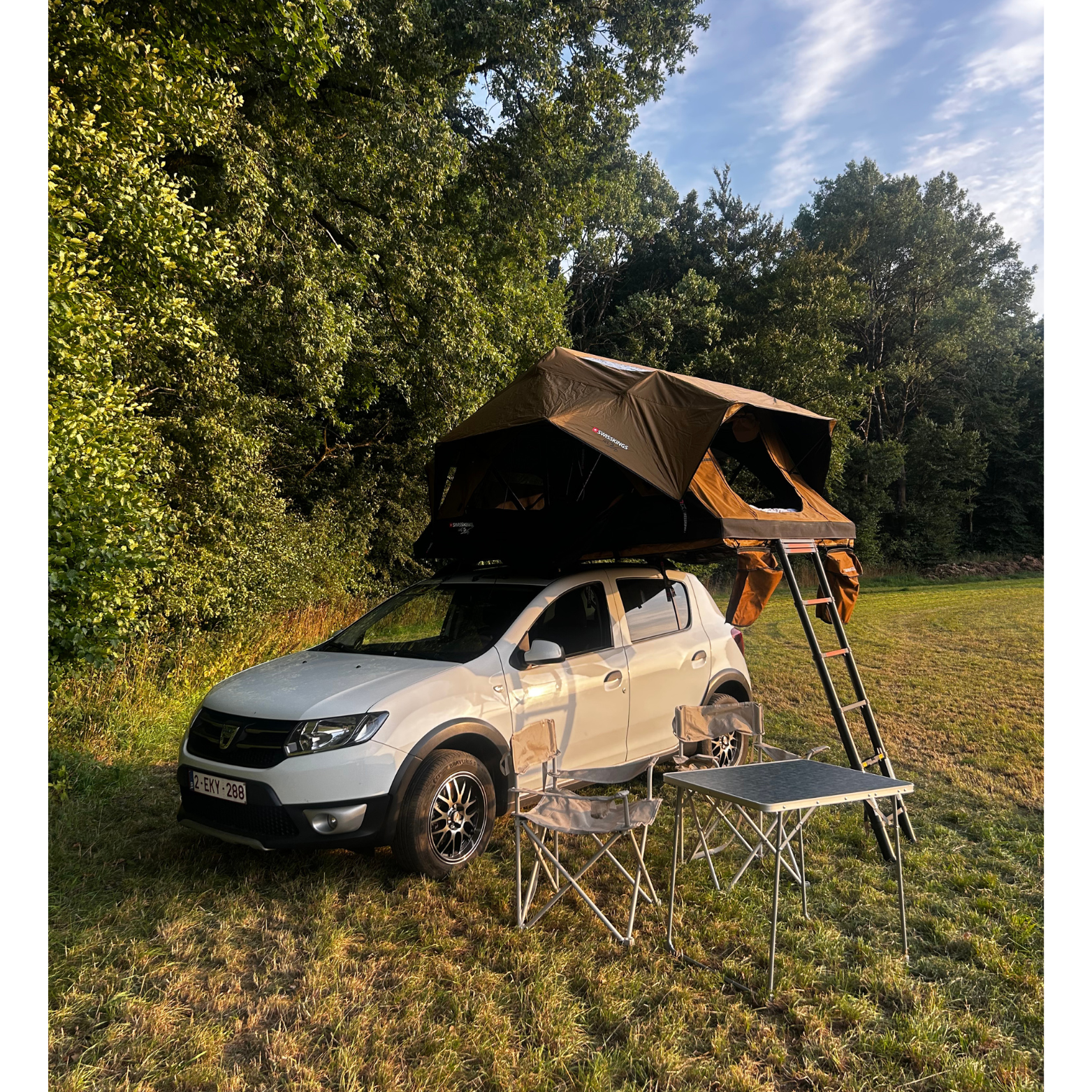 Voiture blanche équipée d'un toit de camping avec tente montée, sur un terrain herbeux avec un fond d'arbres et un ciel bleu, accompagnée d'une petite table pliable et de deux chaises.