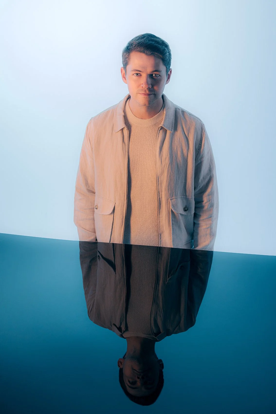 A young man with short brown hair wearing a cream-colored jacket over a beige sweater, standing in front of a light blue background. The lower half of the image reflects his upper body on a glossy black surface.