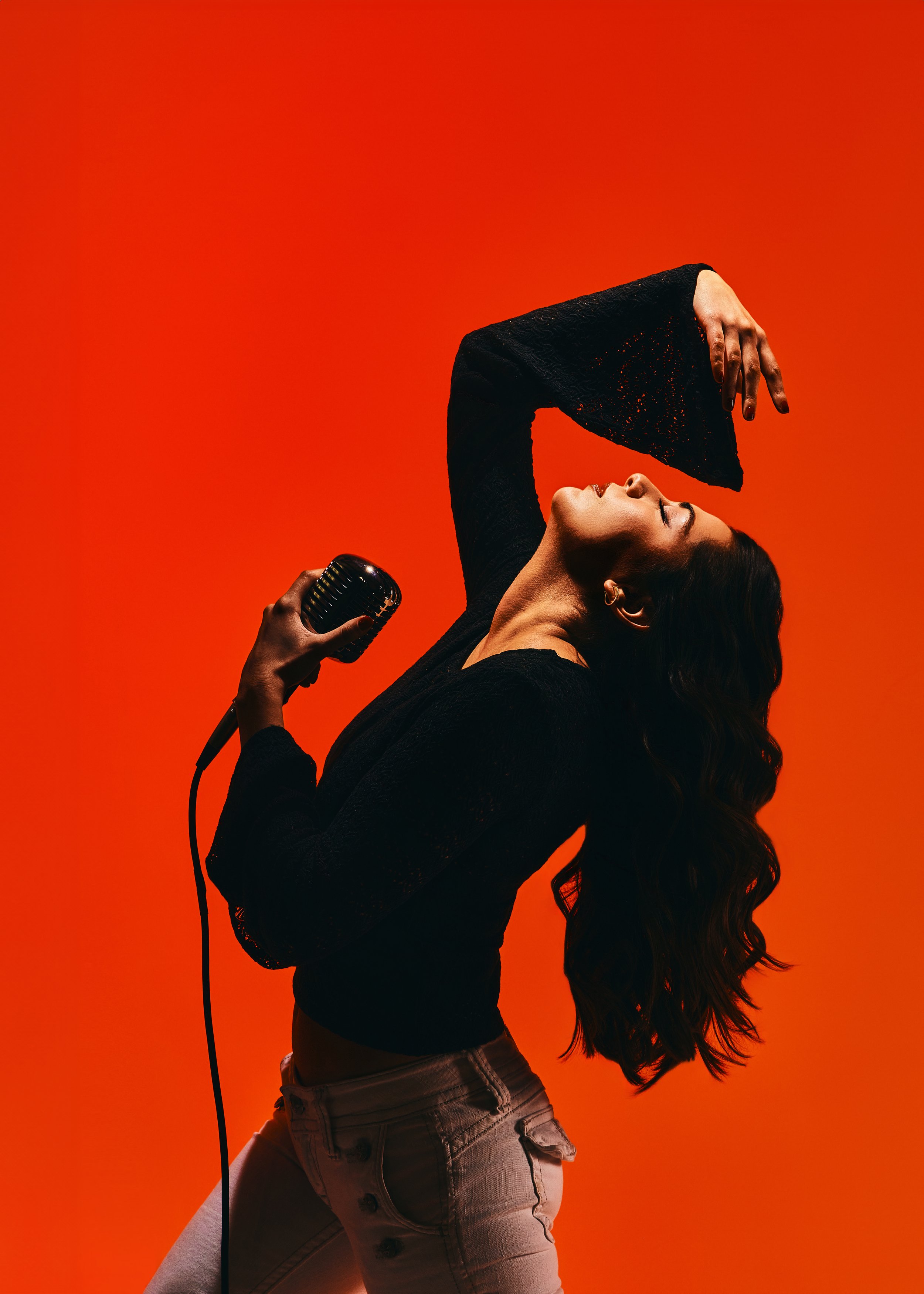 A woman with long, wavy hair is singing or performing into a vintage microphone, with her head tilted back and one arm raised, against an orange background. She is wearing a black long-sleeve top and light-colored pants.