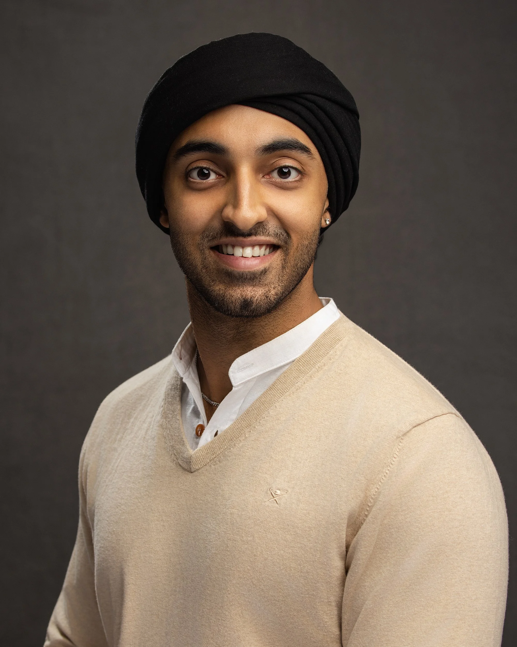A young man smiling with a black turban, wearing a beige sweater over a white collared shirt, against a dark gray background.