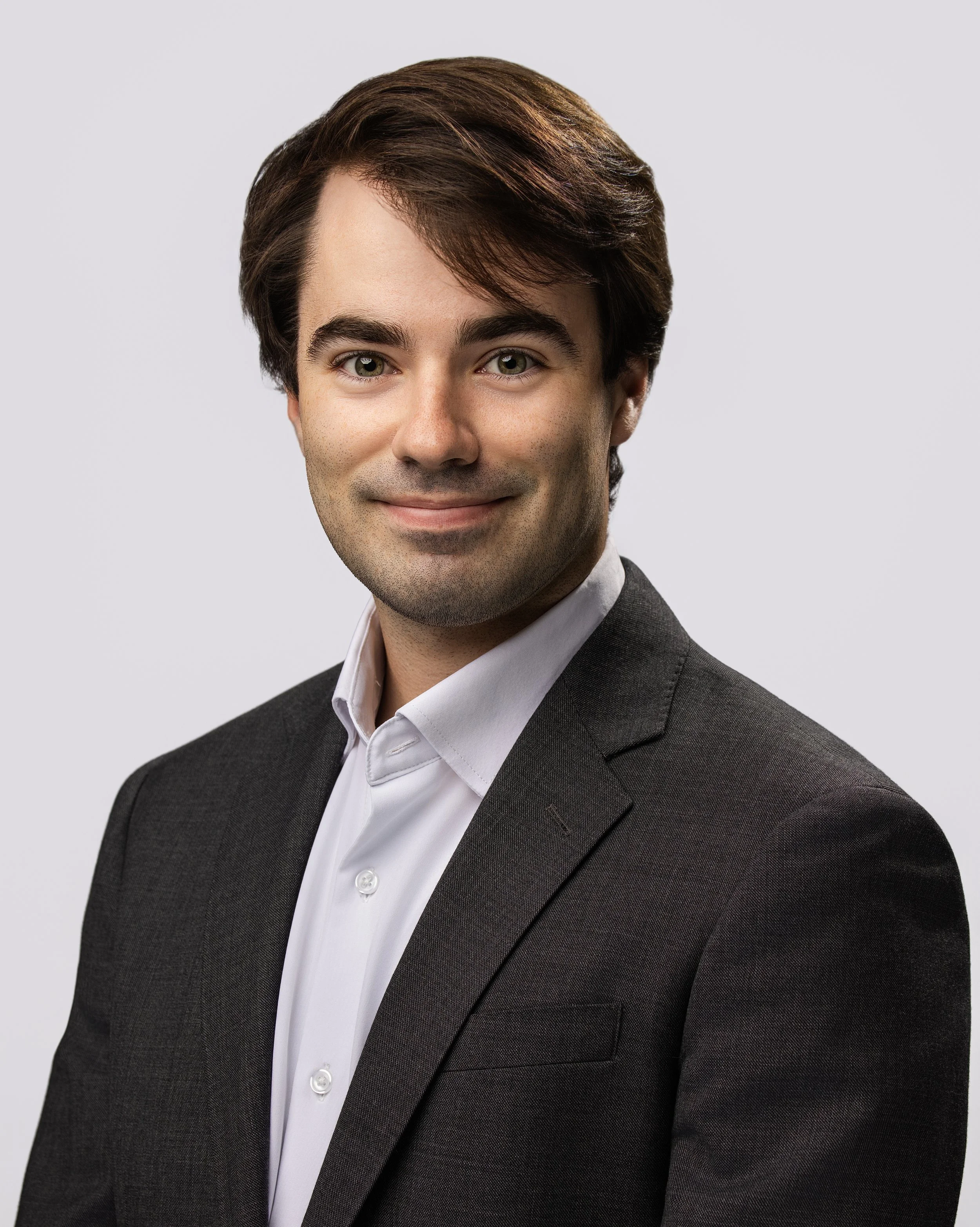 Professional headshot of a young man with brown hair, wearing a dark suit and white shirt, smiling against a light gray background.