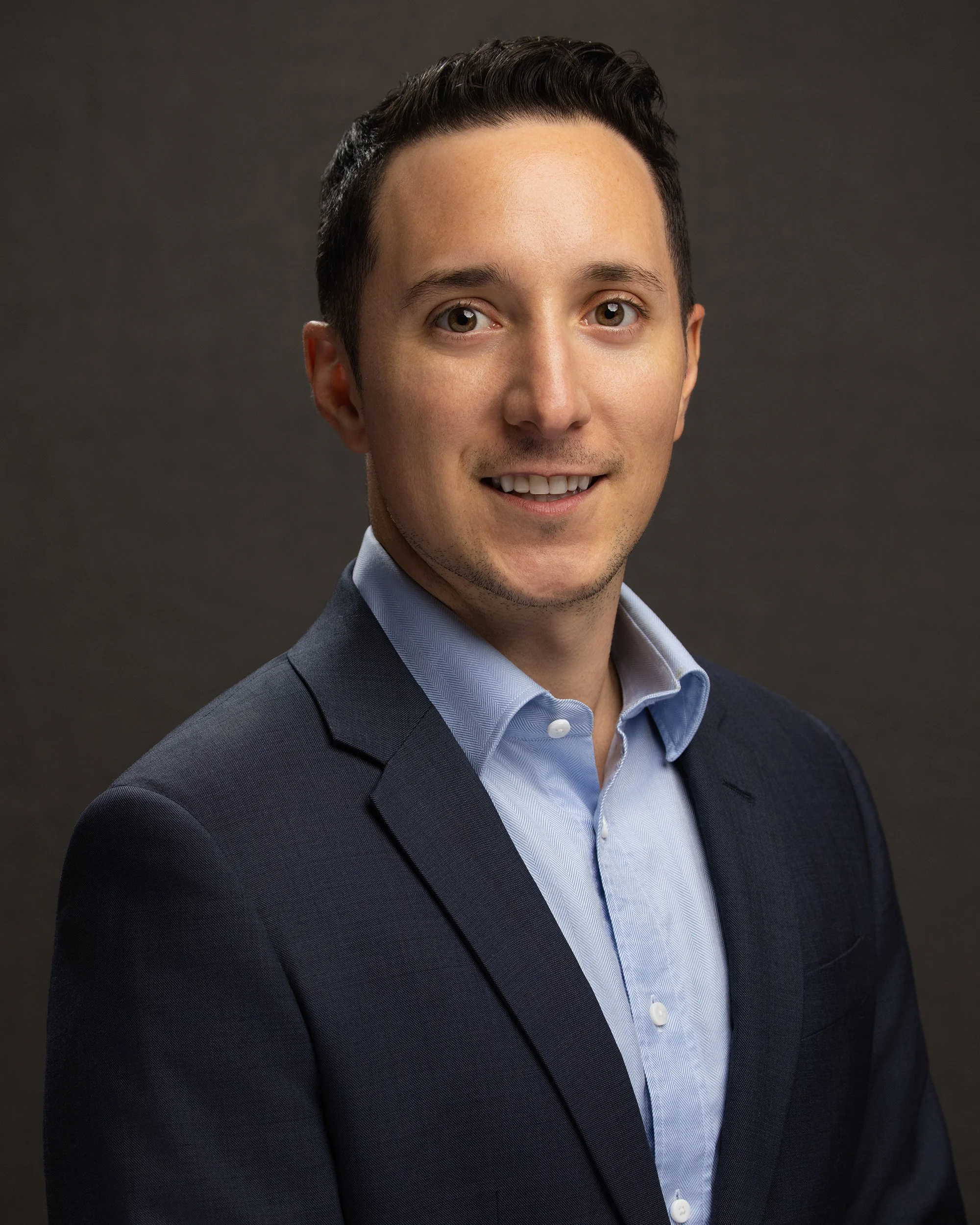 Professional headshot of a smiling man in a dark suit and light blue dress shirt against a gray background.
