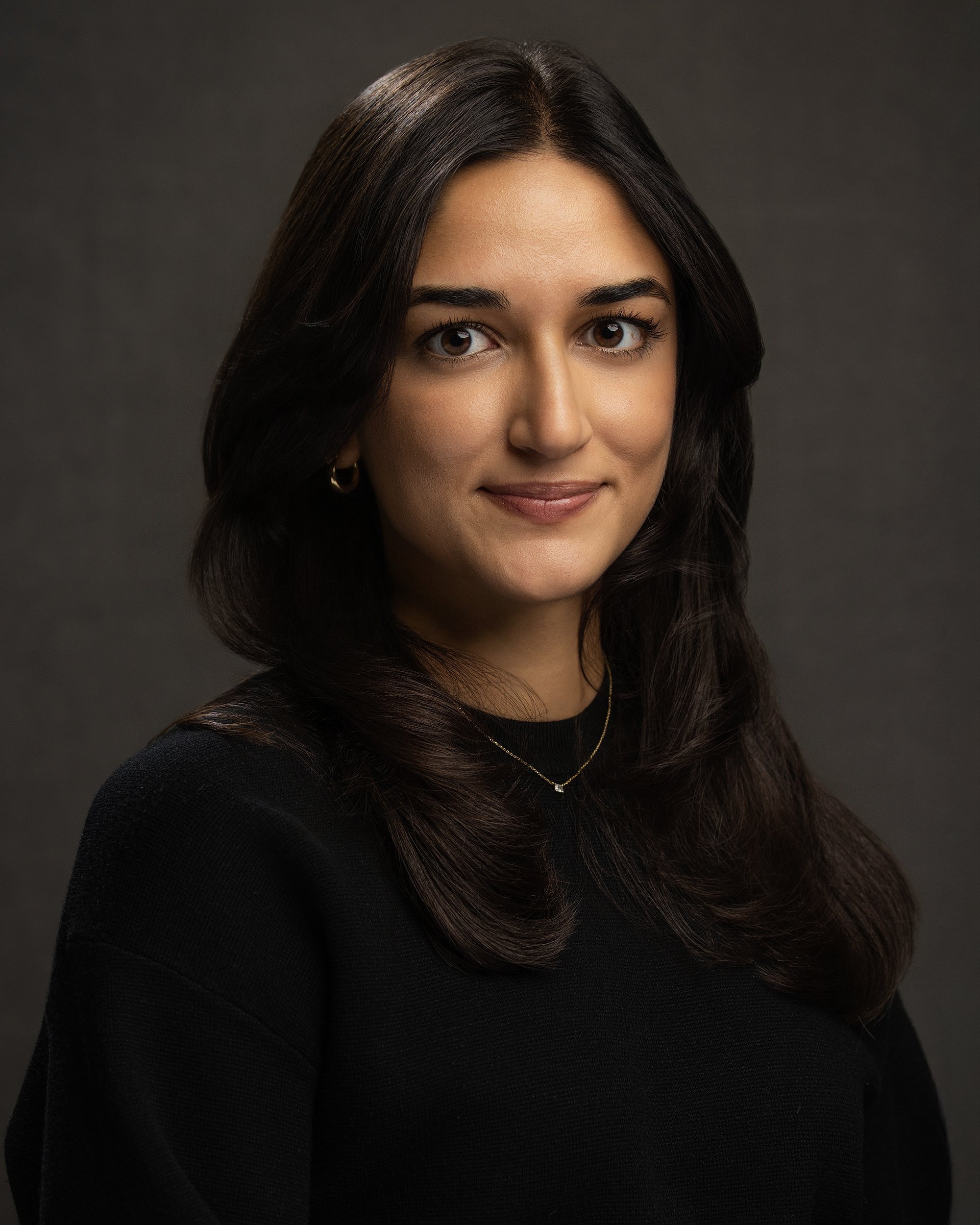 Portrait of a woman with dark hair, wearing a black top, gold hoop earrings, and a delicate necklace, against a dark background.