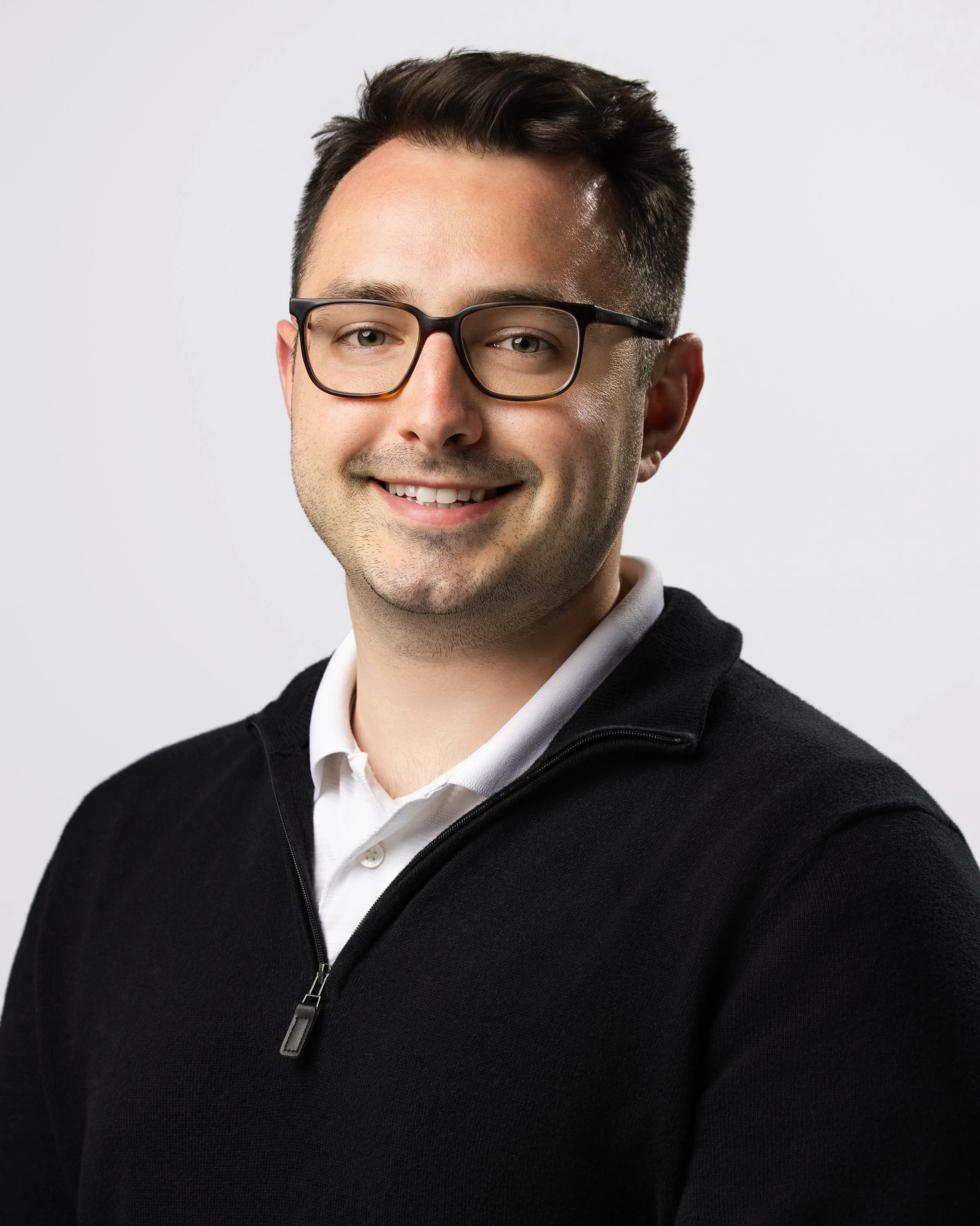 Portrait of a young man wearing glasses, a white collared shirt, and a black zip-up sweater, smiling at the camera against a plain white background.
