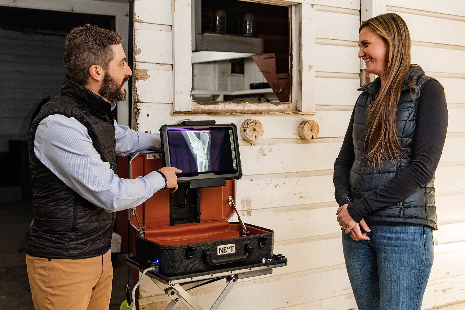 A man showing a digital X-ray of a horse’s leg to a woman in a barn or rustic setting.