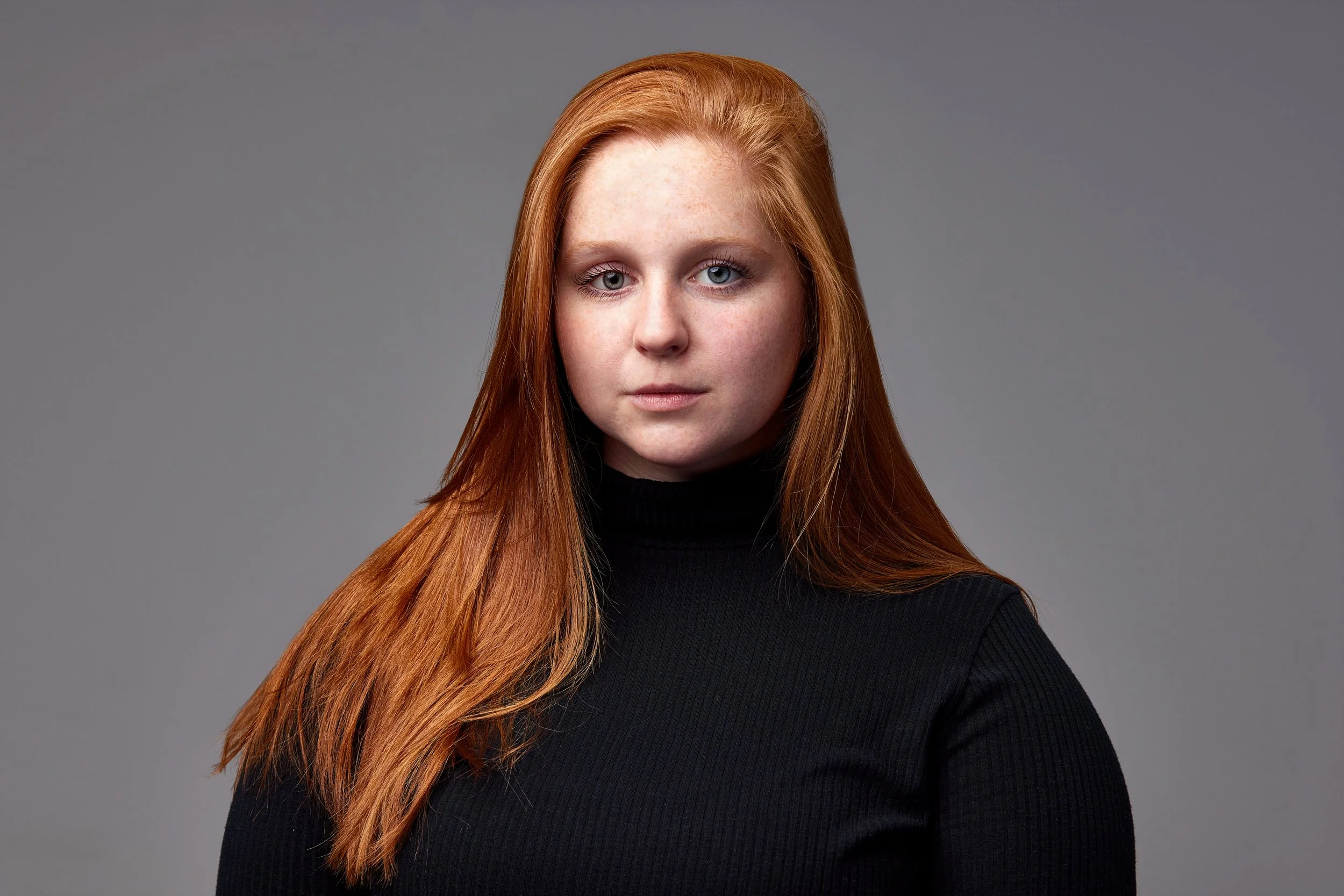 Portrait of a red-haired woman wearing a black turtleneck against a gray background.