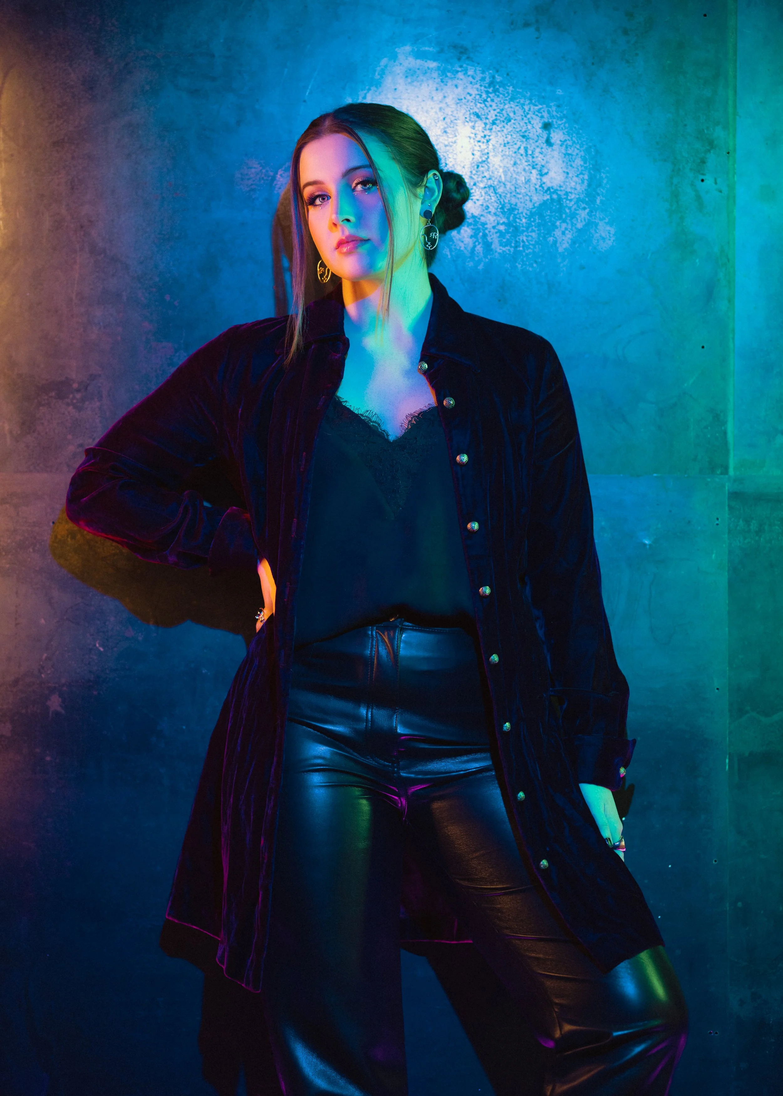 A woman in a black outfit stands against a metallic wall, illuminated by colorful neon lights.