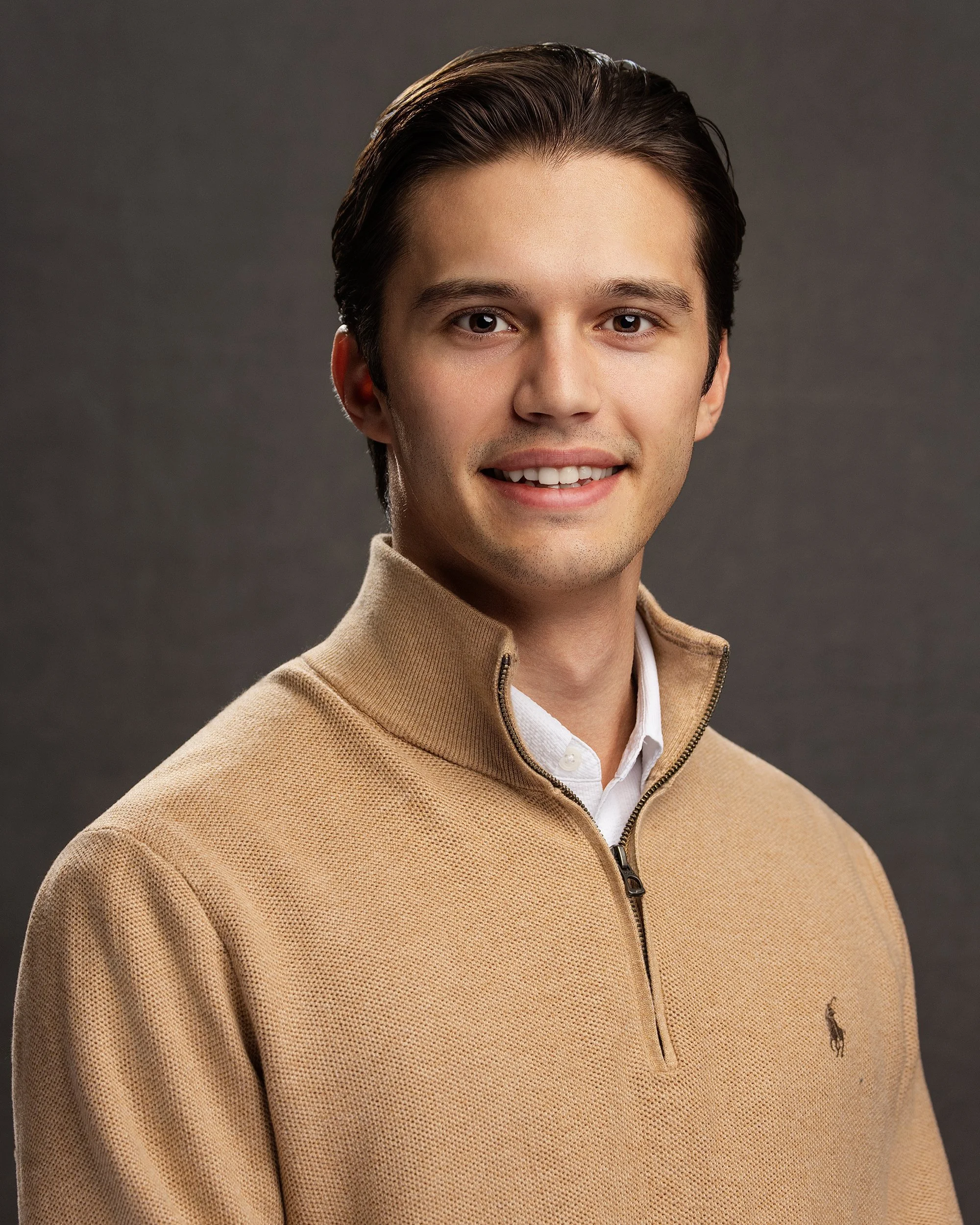 A young man with dark neatly styled hair wearing a beige quarter-zip sweater with a small logo, smiling at the camera against a gray background.