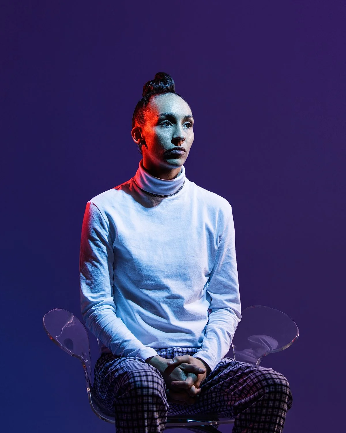 A woman with dark hair tied in a bun, sitting on a transparent chair against a dark background with purple and blue lighting, wearing a white turtleneck and plaid pants.