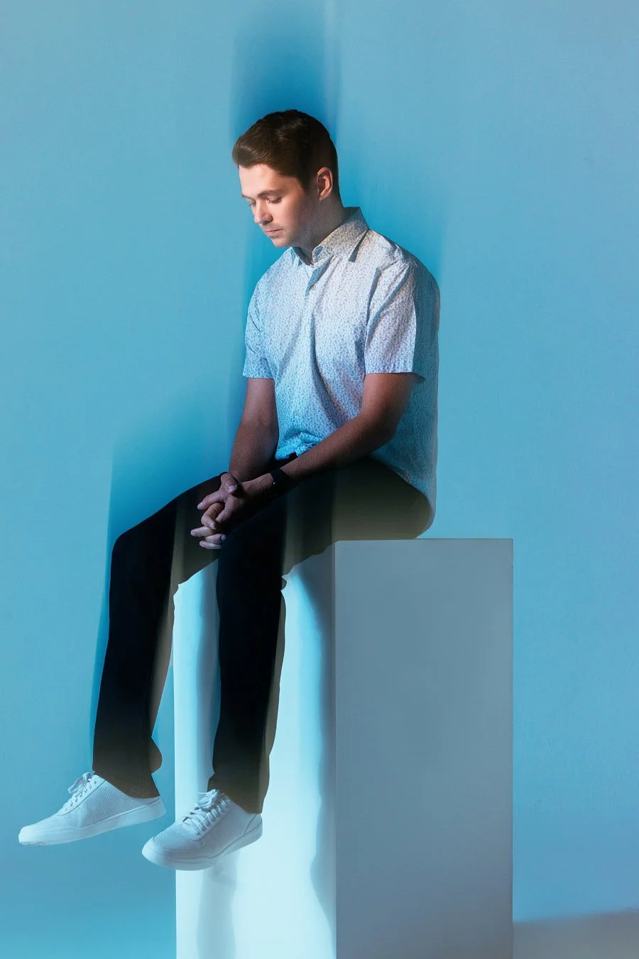 Young man seated on a white block, with hands clasped and head bowed, in a room with cool blue lighting.