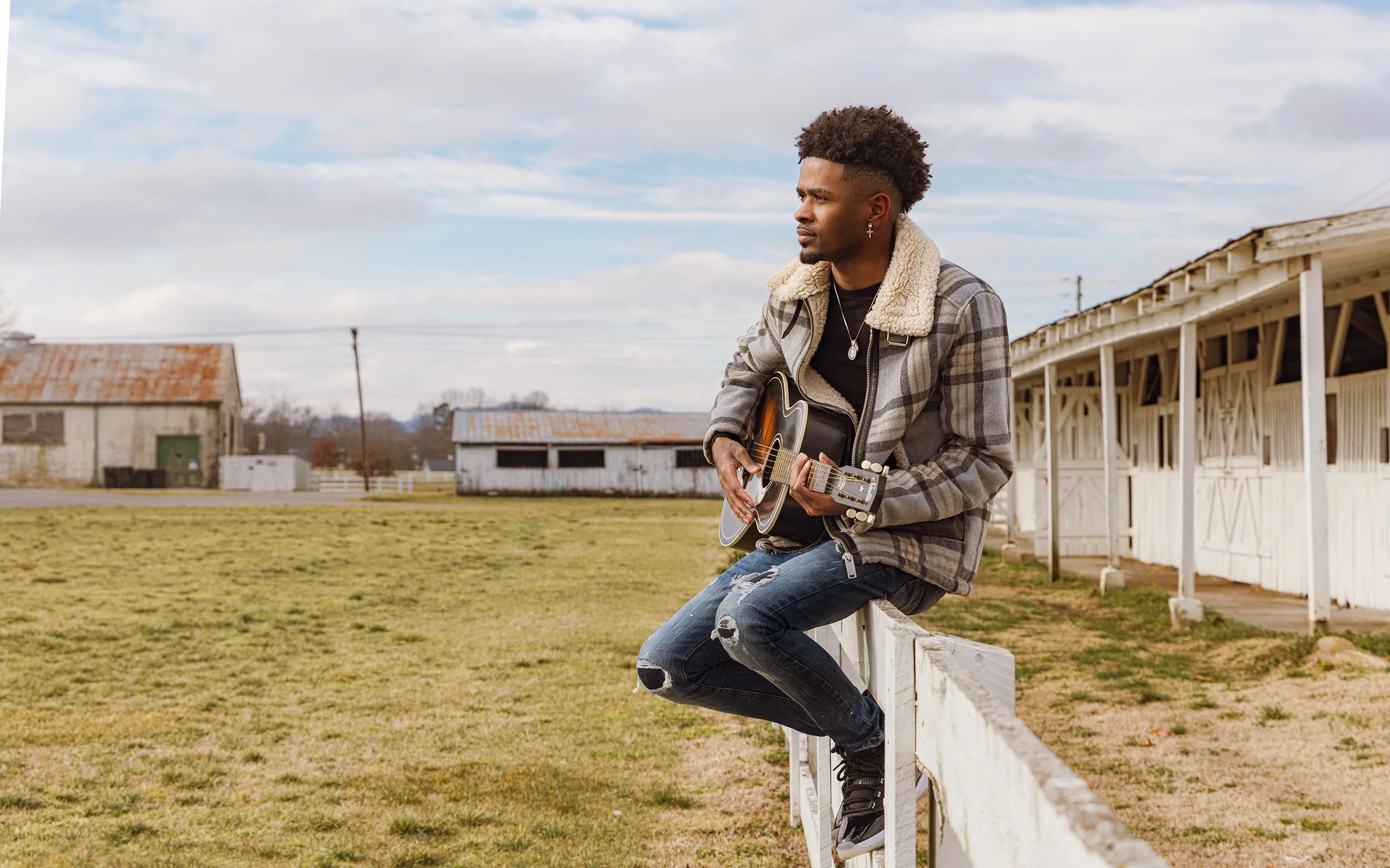 A young man with an afro hairstyle, wearing a plaid coat with a fur collar, black t-shirt, ripped jeans, and sneakers, sits on a white fence playing an acoustic guitar in a rural area with old barns and a partly cloudy sky.