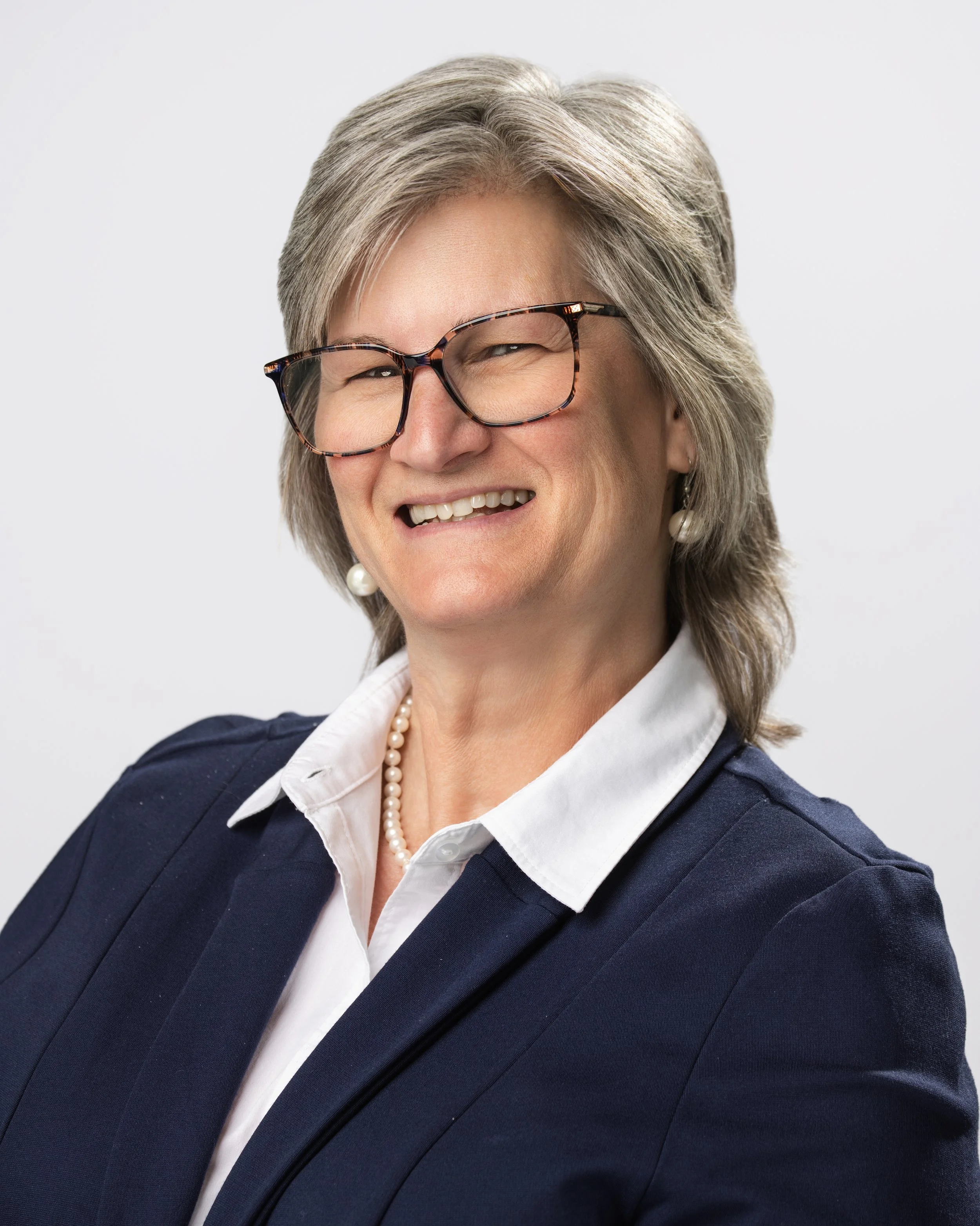 Portrait of a smiling middle-aged woman with short gray hair, glasses, pearl earrings, a pearl necklace, white shirt, and navy blazer against a plain white background.