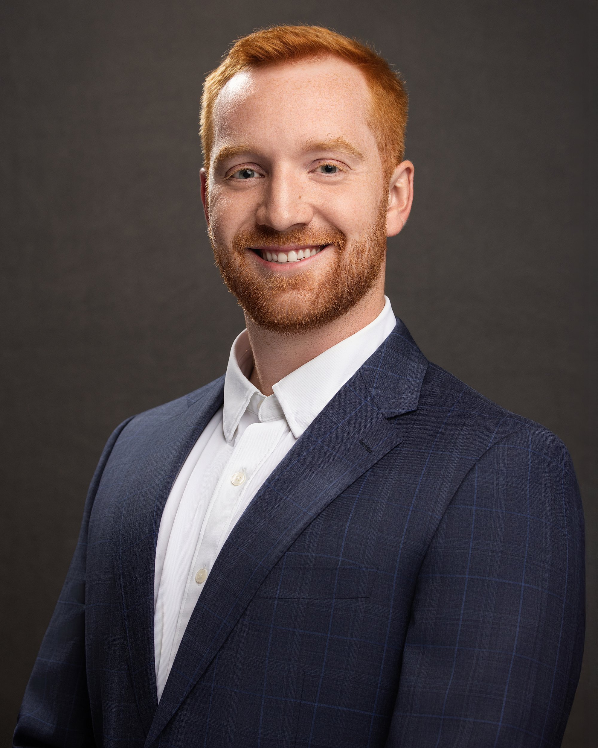Headshot of a smiling man with red hair and beard, dressed in a dark blue checkered suit and white shirt, against a gray background.