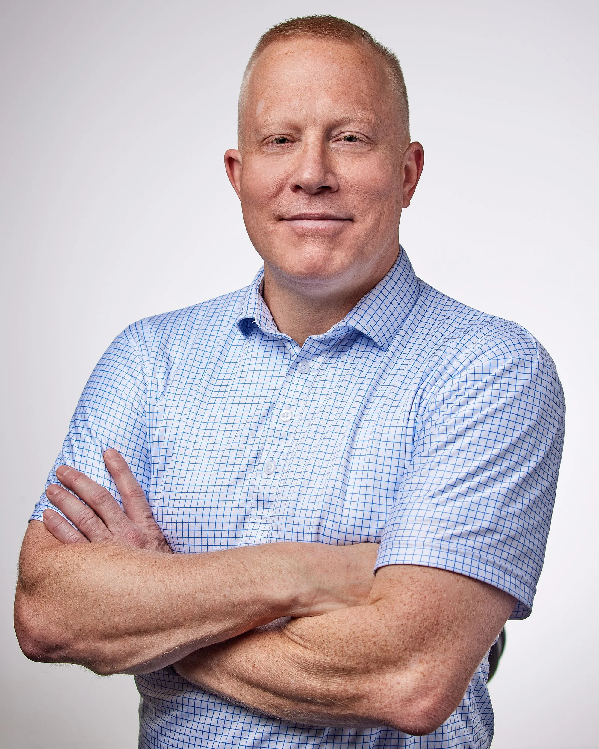 A man with short red hair, wearing a light blue checkered shirt, standing with his arms crossed, smiling slightly, against a plain white background.