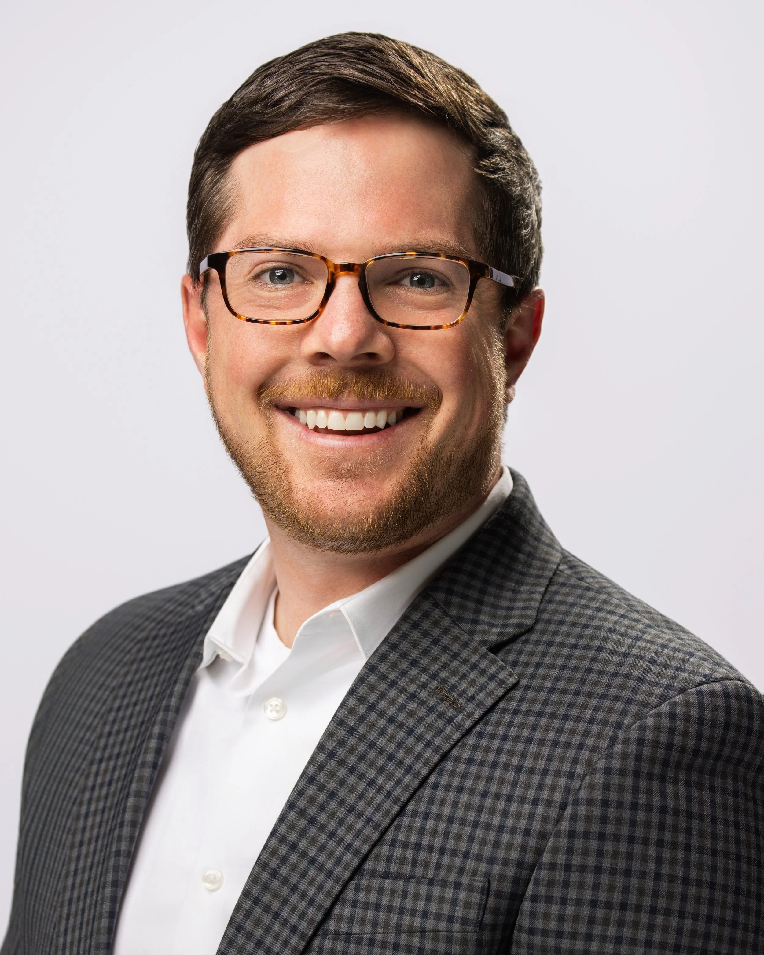Headshot of a smiling man with brown hair, beard, and glasses, wearing a plaid blazer and white shirt against a plain light background.