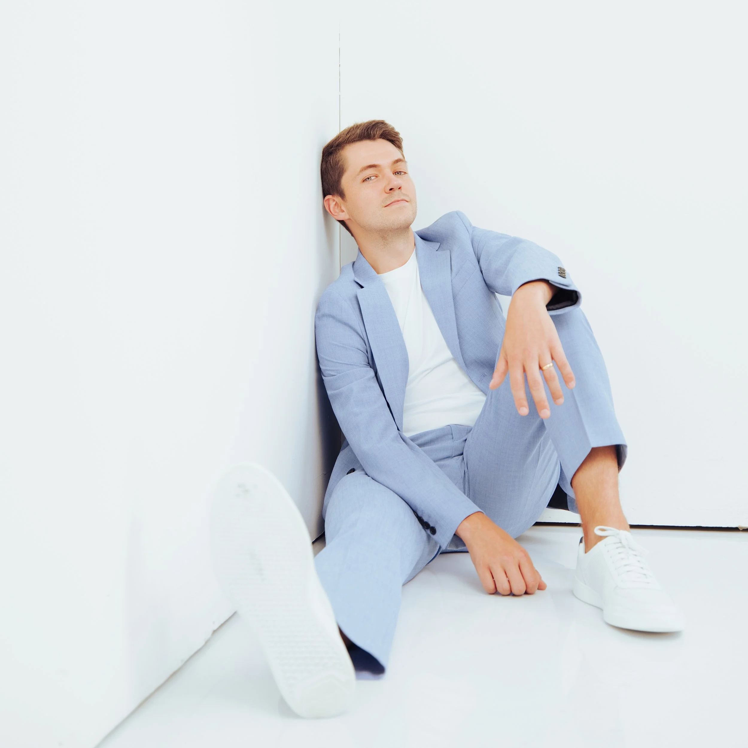 A young man in a light blue suit sitting on the floor in a white room, looking confident.