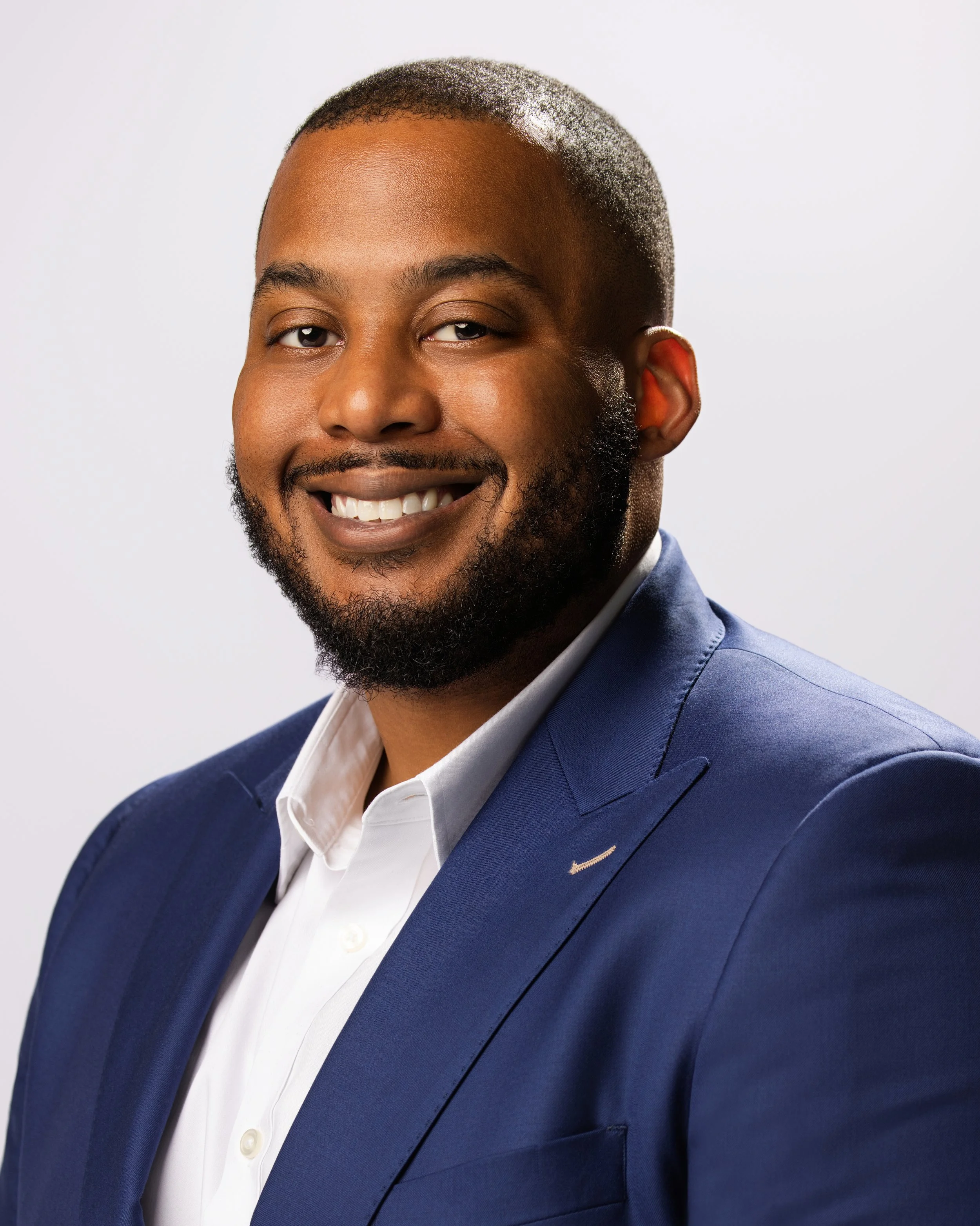 Professional portrait of a smiling African American man in a blue suit and white shirt against a plain background.
