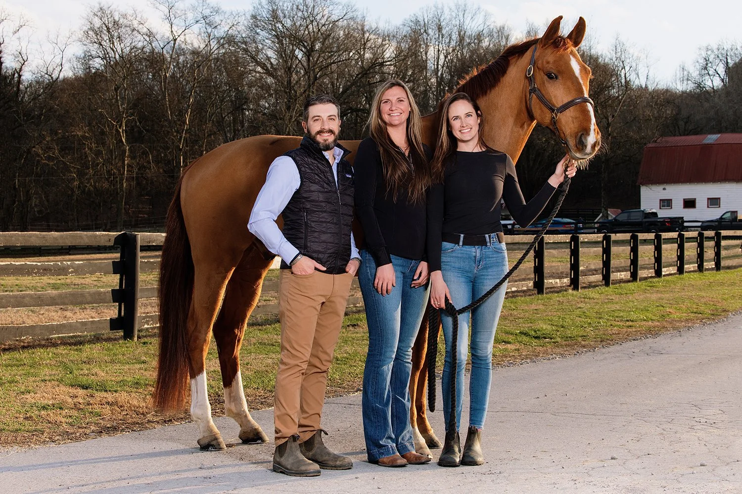 Three people standing with a horse outdoors near a fencing and trees in the background, during daytime.