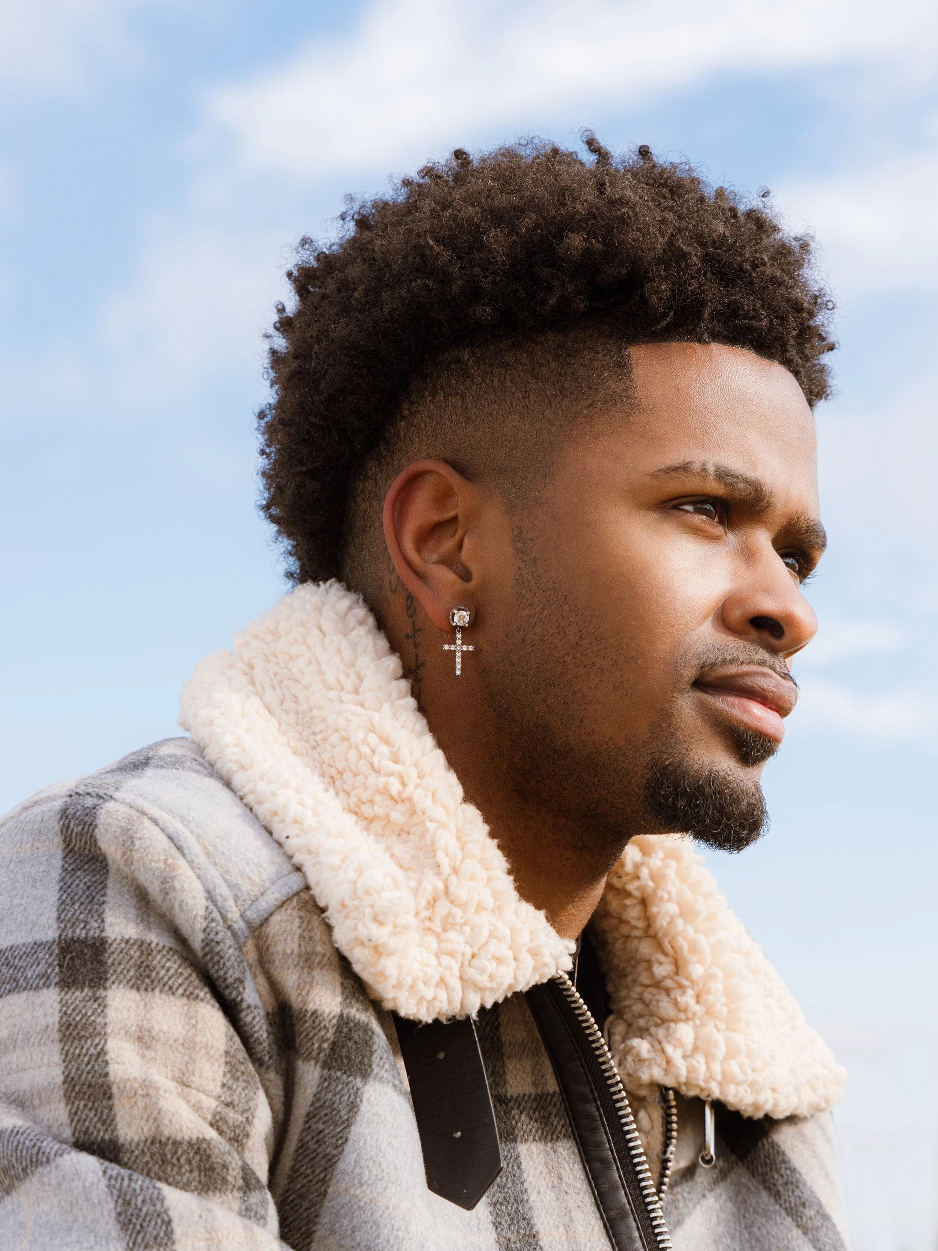 A young man with curly hair and an earring, wearing a plaid jacket with a sherpa collar, looking towards the sky against a cloudy blue sky.