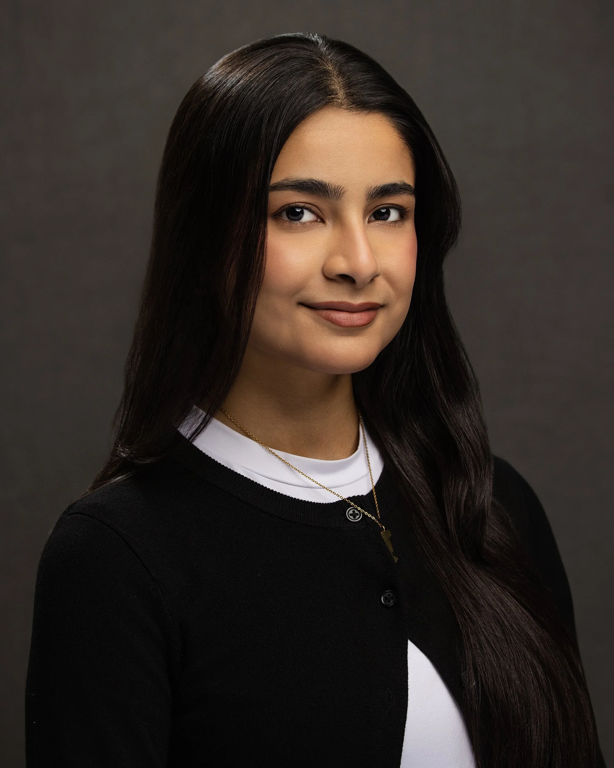 A woman with long dark hair, wearing a black cardigan over a white shirt, with gold necklaces, smiling against a dark gray background.
