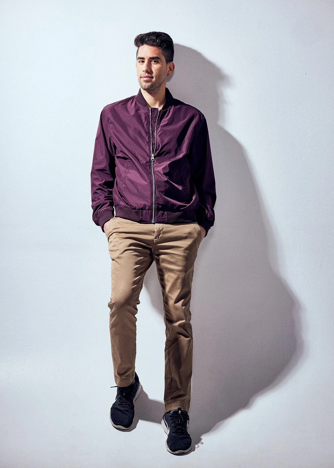 A young man with dark hair, wearing a purple bomber jacket, beige pants, and black sneakers, standing against a plain white wall with his hands in his pockets.