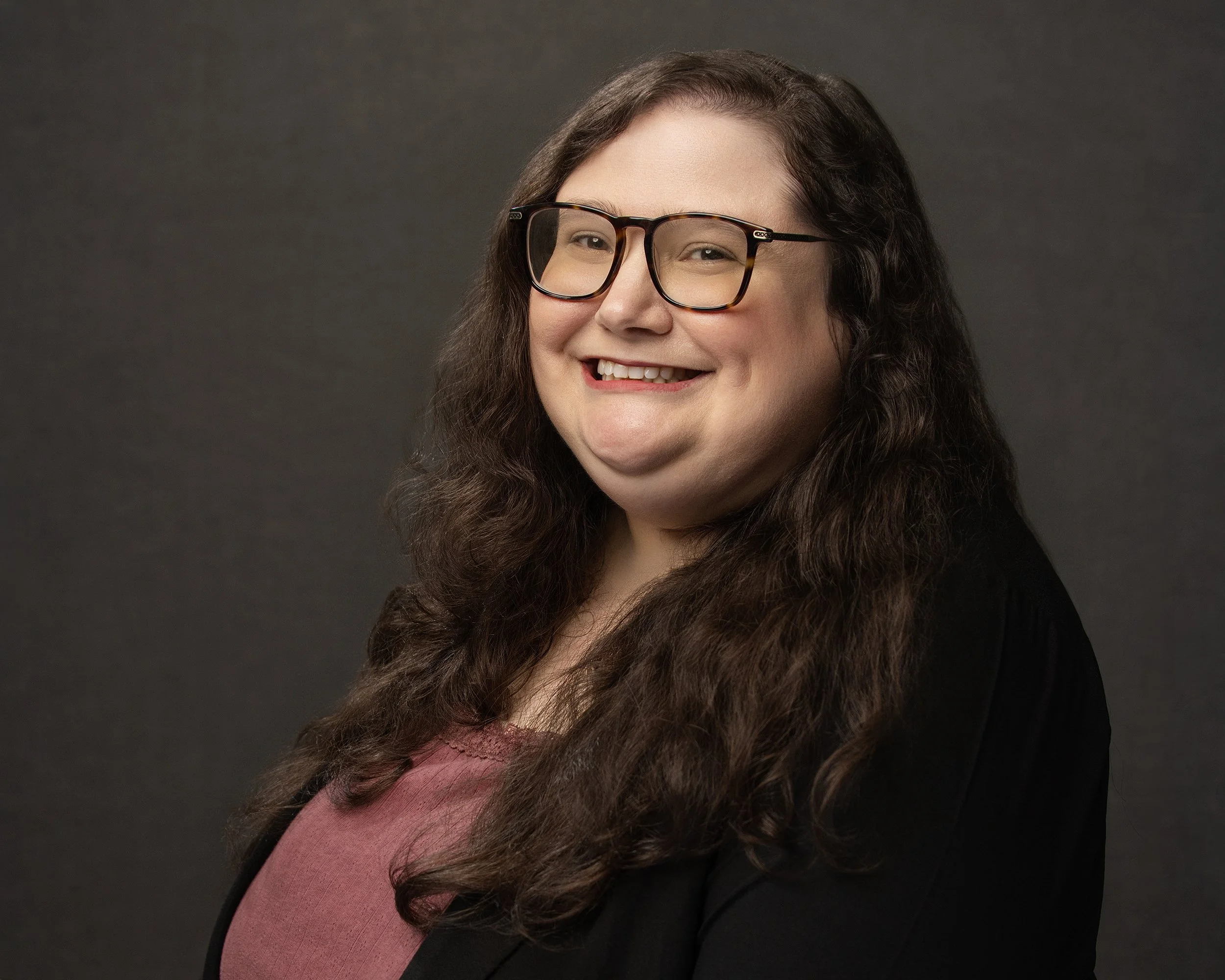 A woman with long, curly brown hair and glasses smiling at the camera, wearing a black jacket and a mauve top, against a dark gray background.