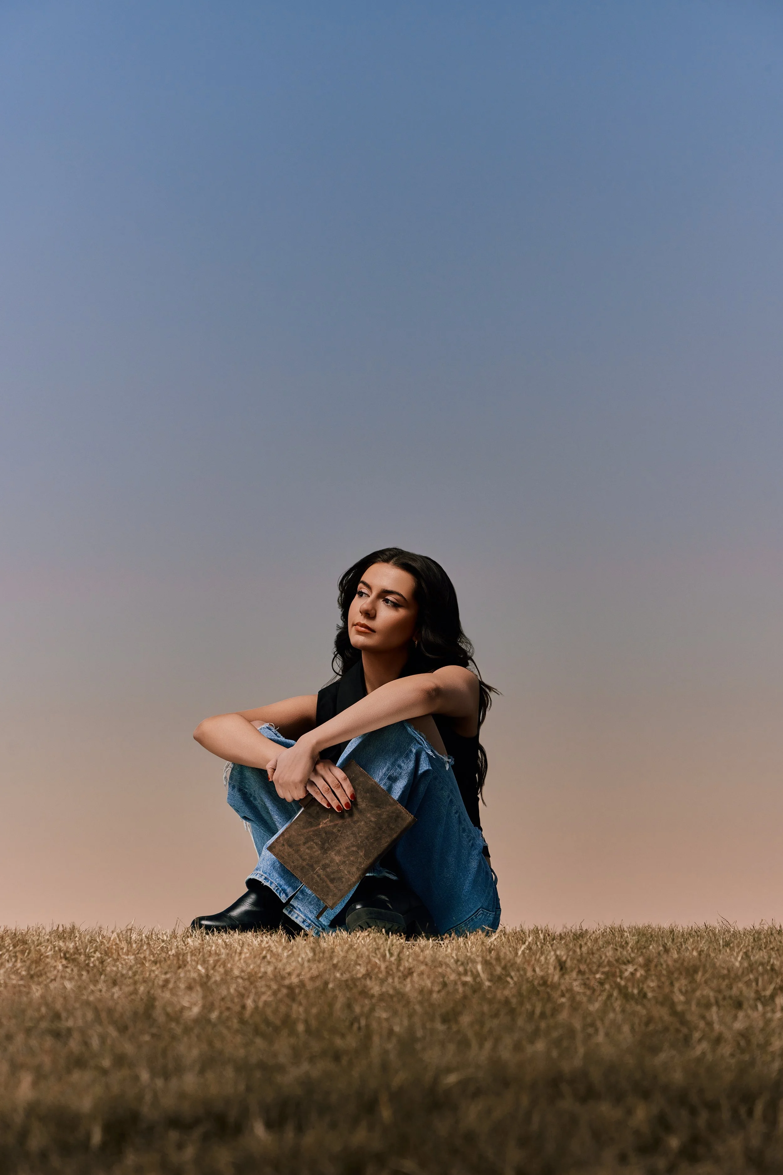 A young woman with long dark hair sitting on grass, holding a book, with a gradient sunset sky in the background.