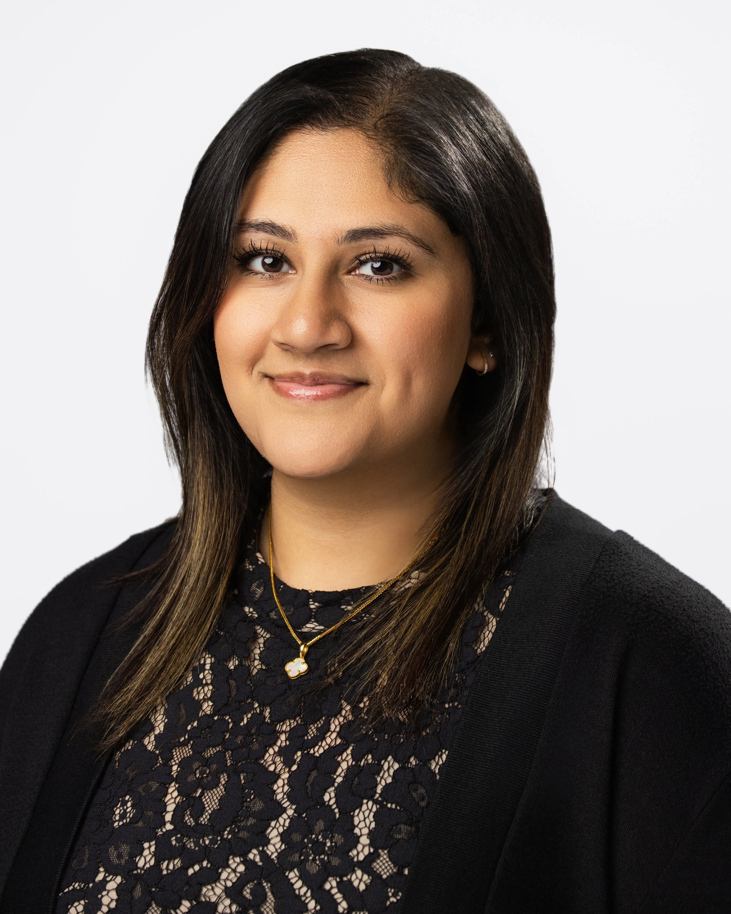 Professional headshot of a woman with shoulder-length dark hair, wearing a black lace top and a gold necklace with a white flower pendant, smiling softly against a white background.