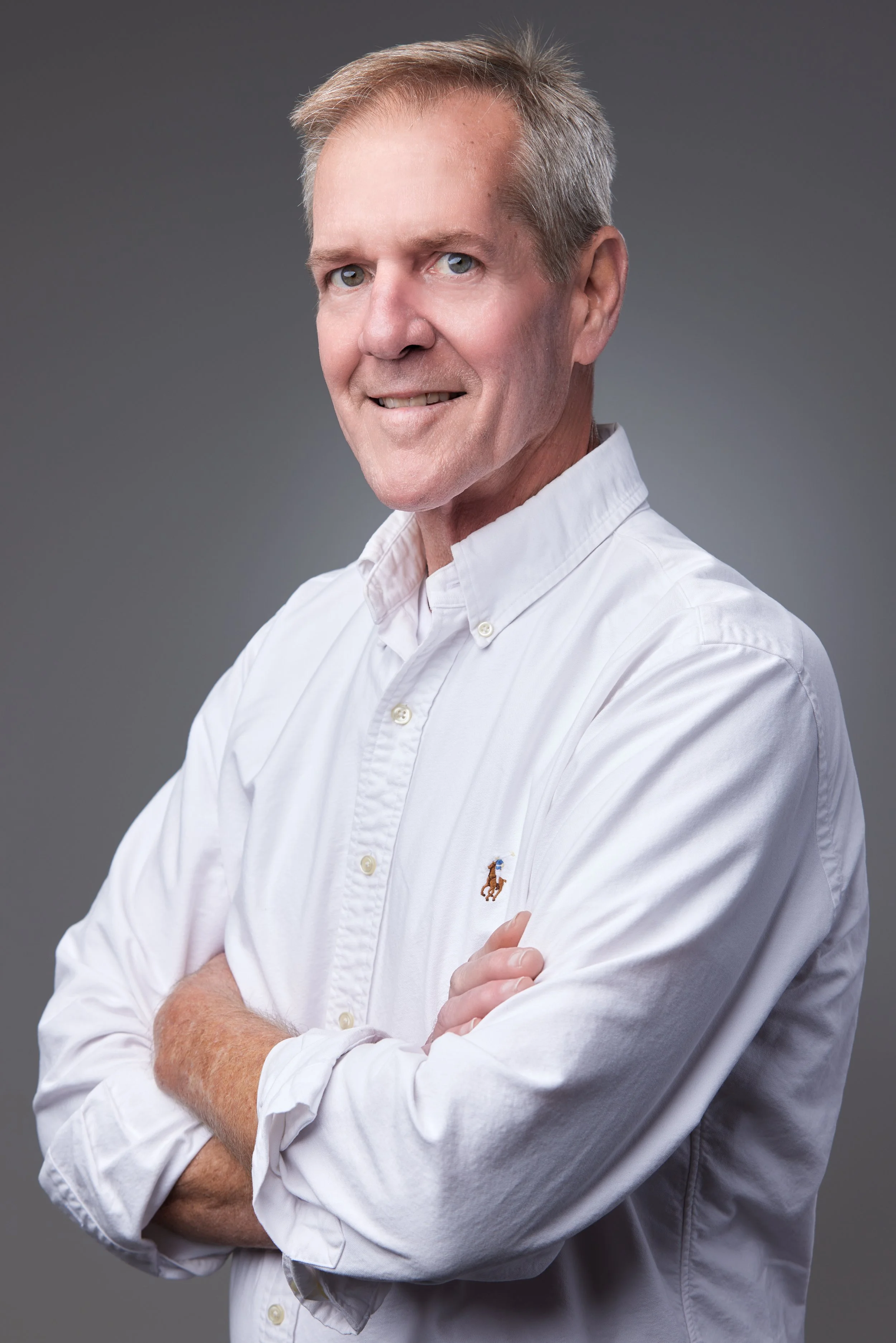 A middle-aged man with short, light-colored hair wearing a white button-up shirt with a Polo Ralph Lauren logo, standing with arms crossed against a gray background.