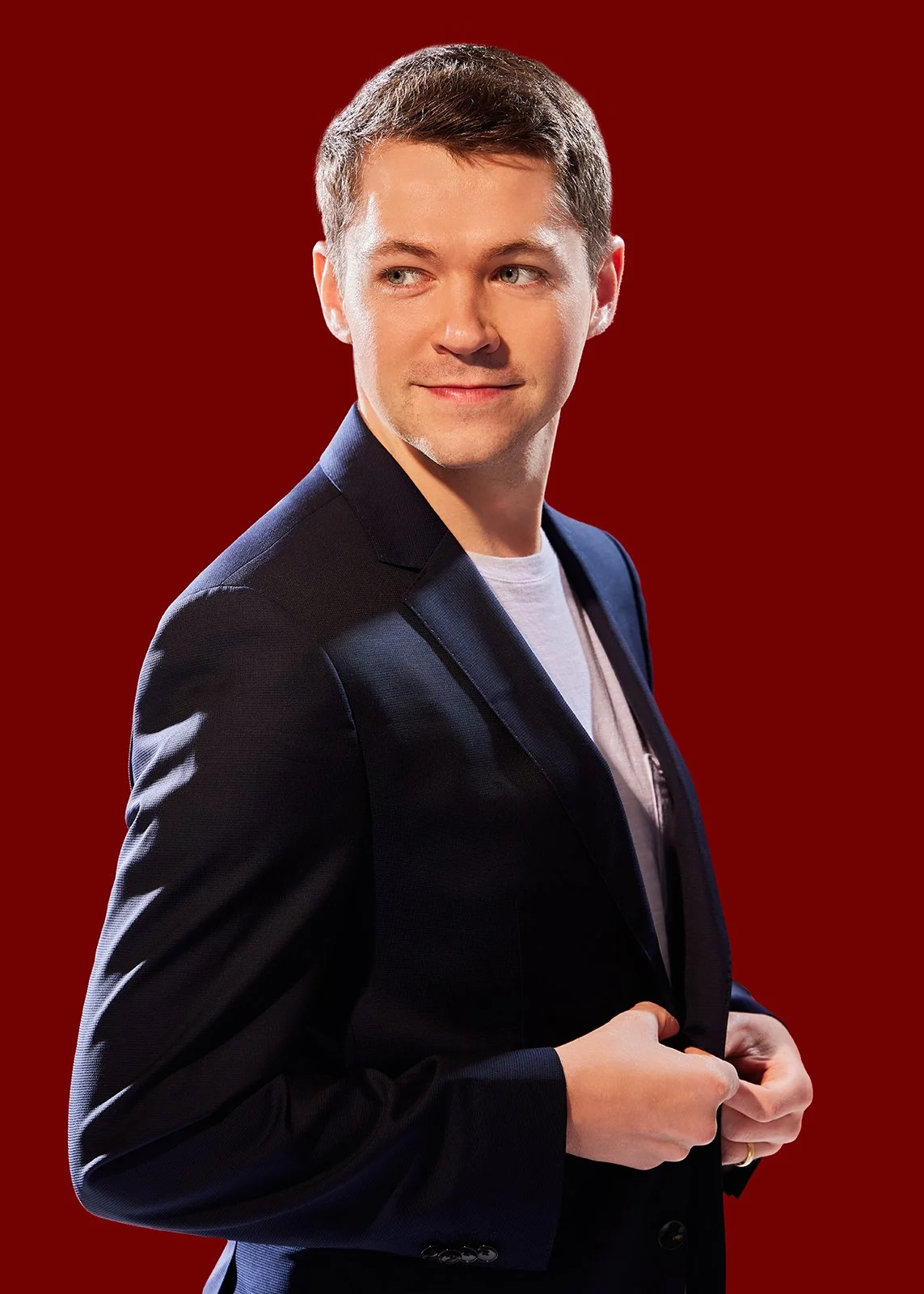 A young man with short brown hair, wearing a dark blazer over a white t-shirt, is adjusting his blazer while looking to the side against a red background.
