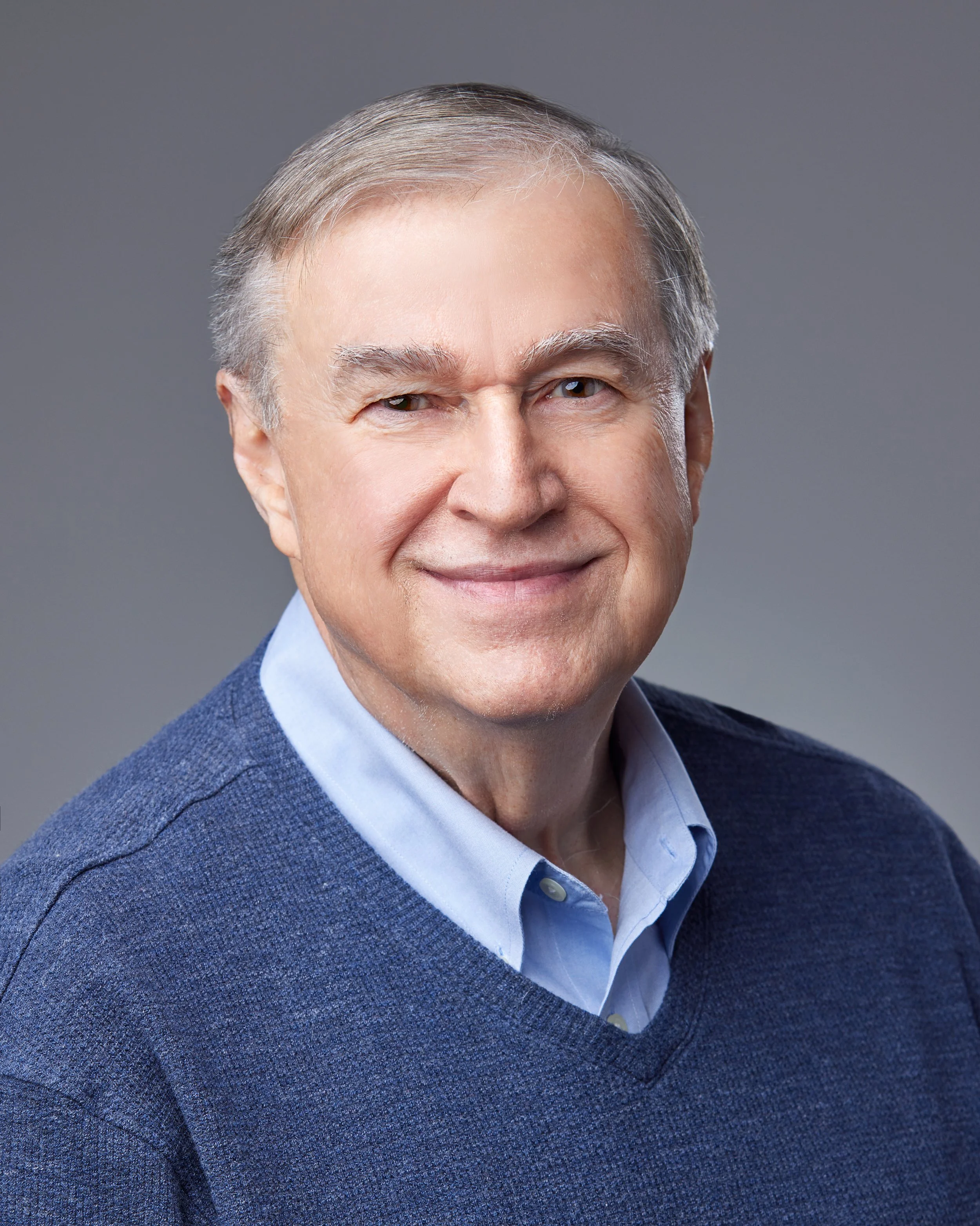Portrait of an older man with gray hair, smiling, wearing a blue sweater over a collared shirt, against a gray background.