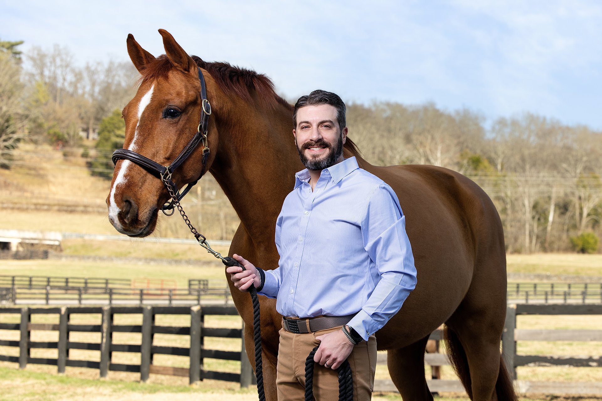 A man with a beard and blue dress shirt holding a chestnut horse with a black halter in an outdoor farm setting.