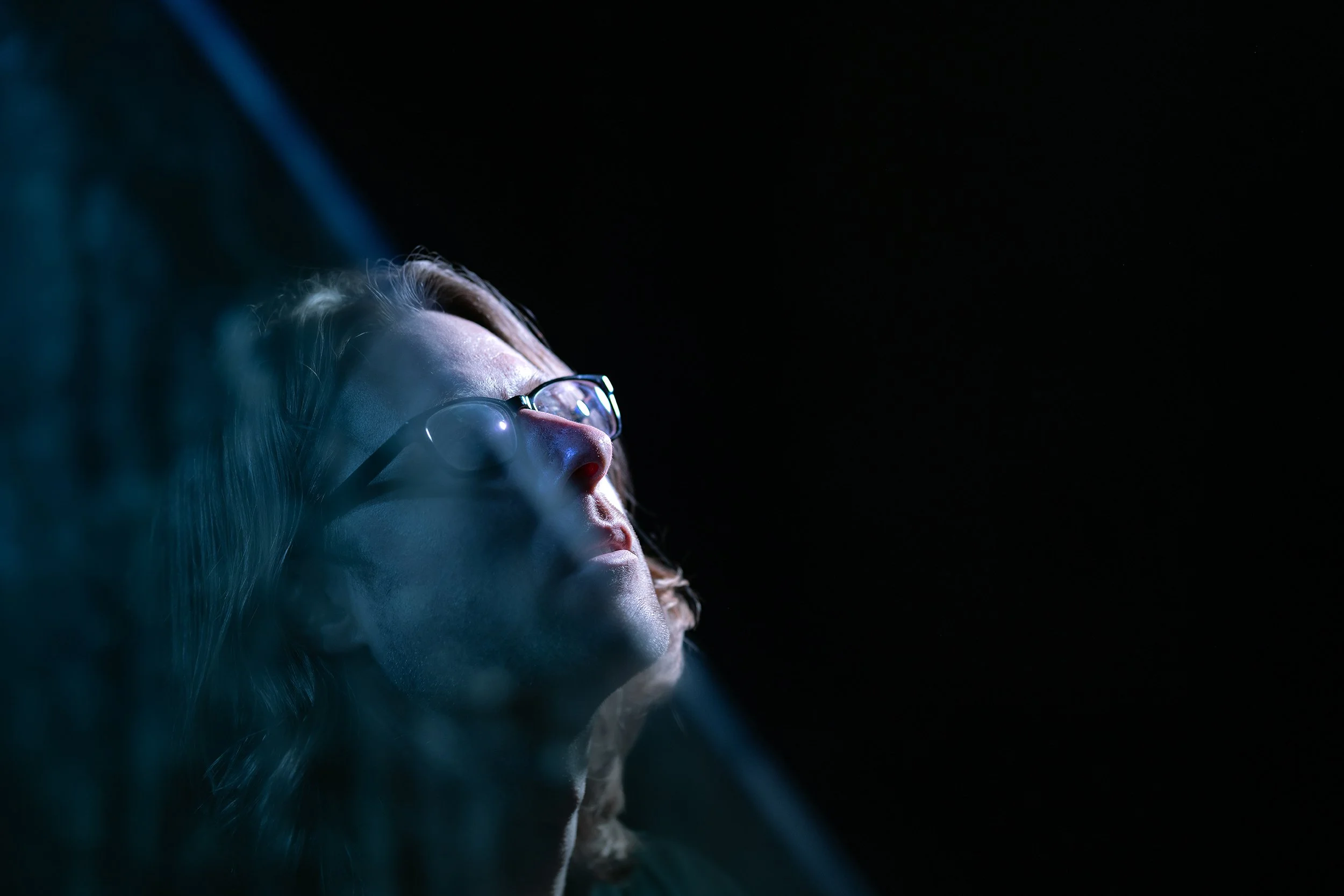 A woman with glasses and closed eyes illuminated by soft light on her face against a dark background.