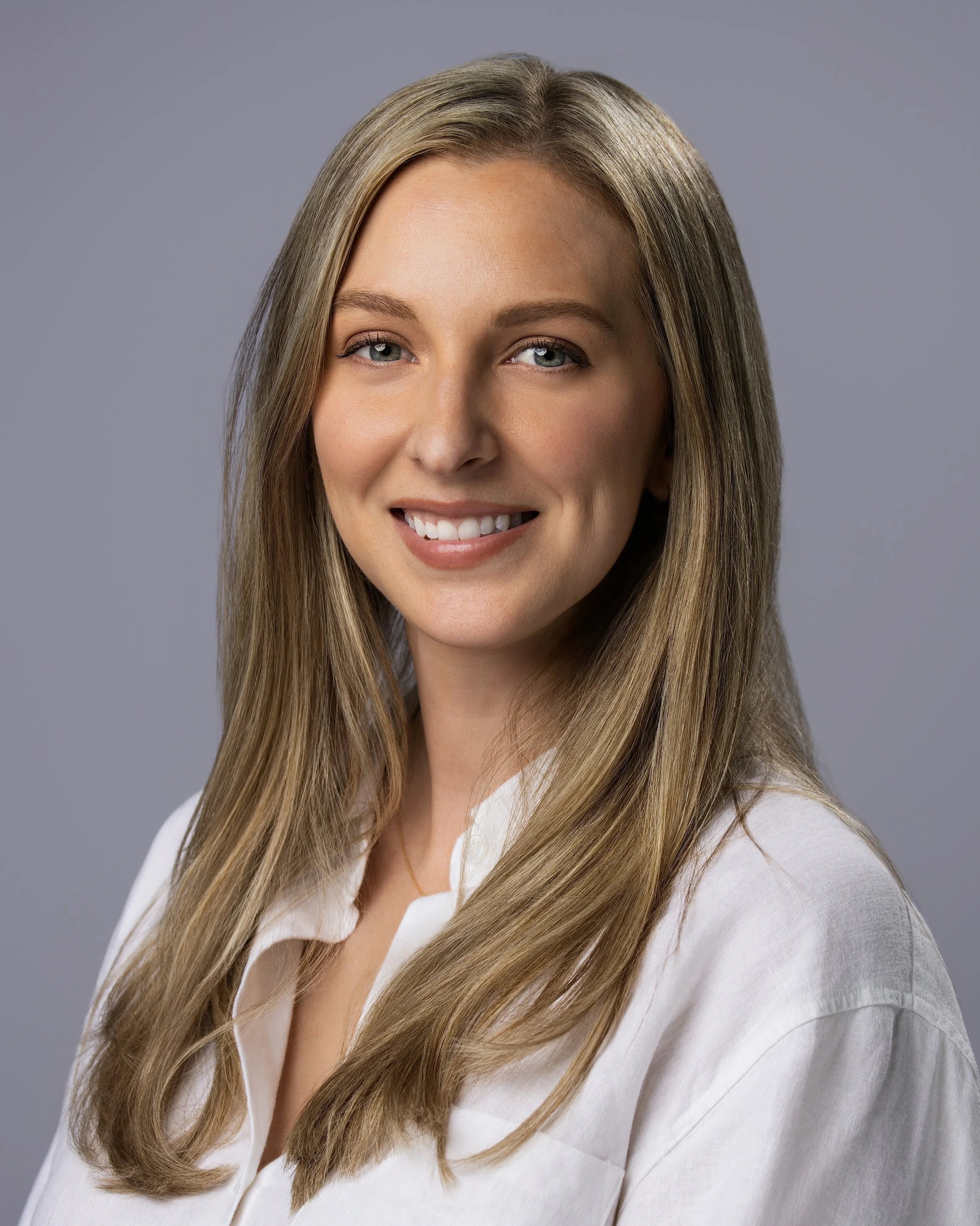 A smiling woman with long, straight blonde hair, wearing a white blouse, against a plain gray background.