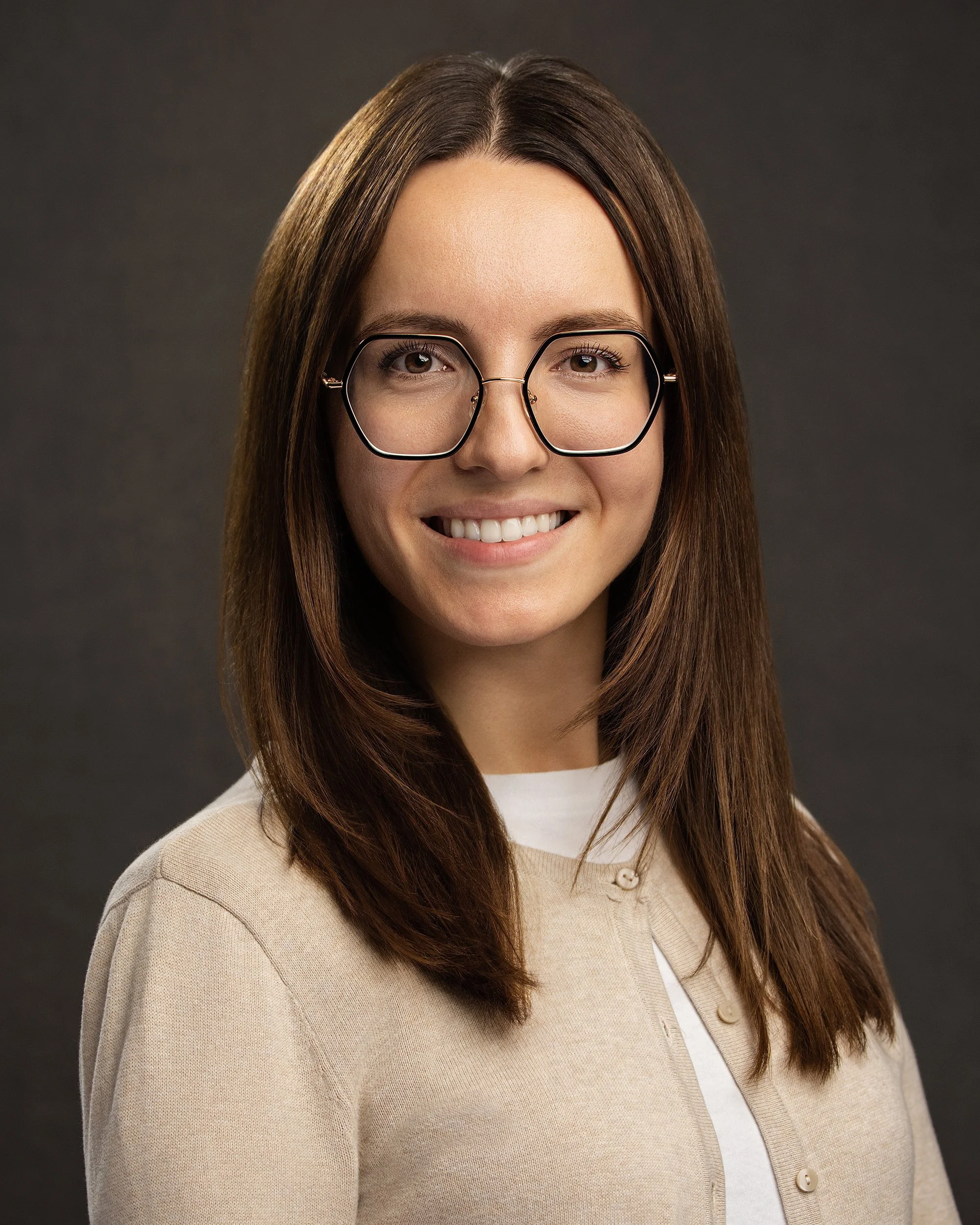 A young woman with long brown hair and glasses, smiling, wearing a beige cardigan over a white shirt, against a dark gray background.
