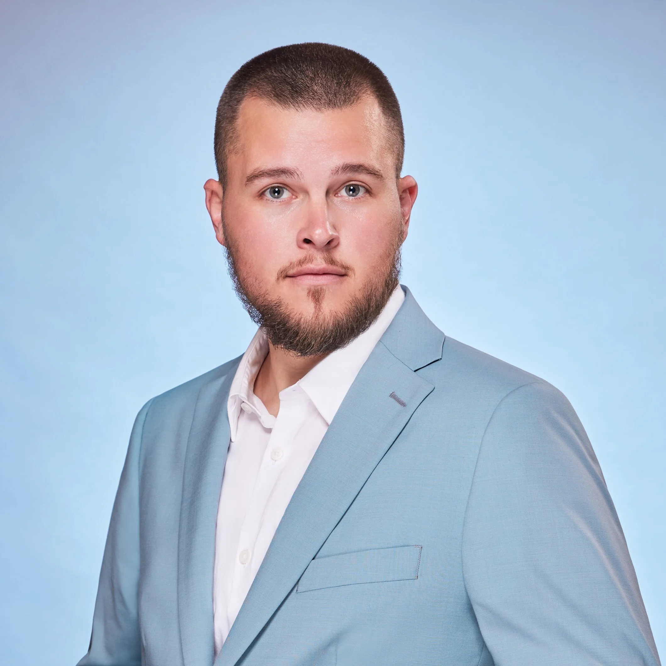 Headshot of a young man with short hair and a beard, wearing a light blue suit and white shirt against a light blue background.