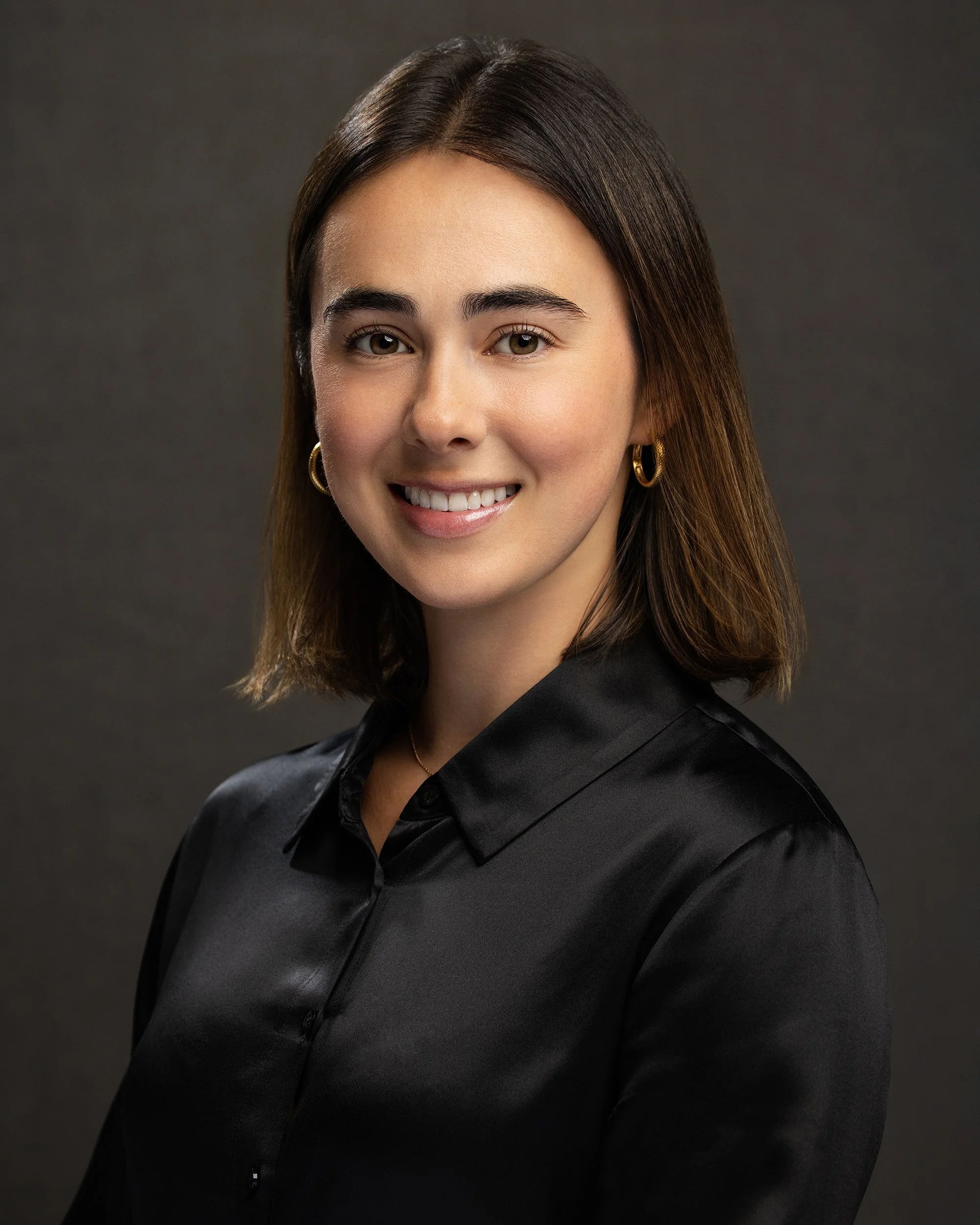 A woman with shoulder-length brown hair, wearing a black satin shirt and gold hoop earrings, smiling at the camera against a dark background.