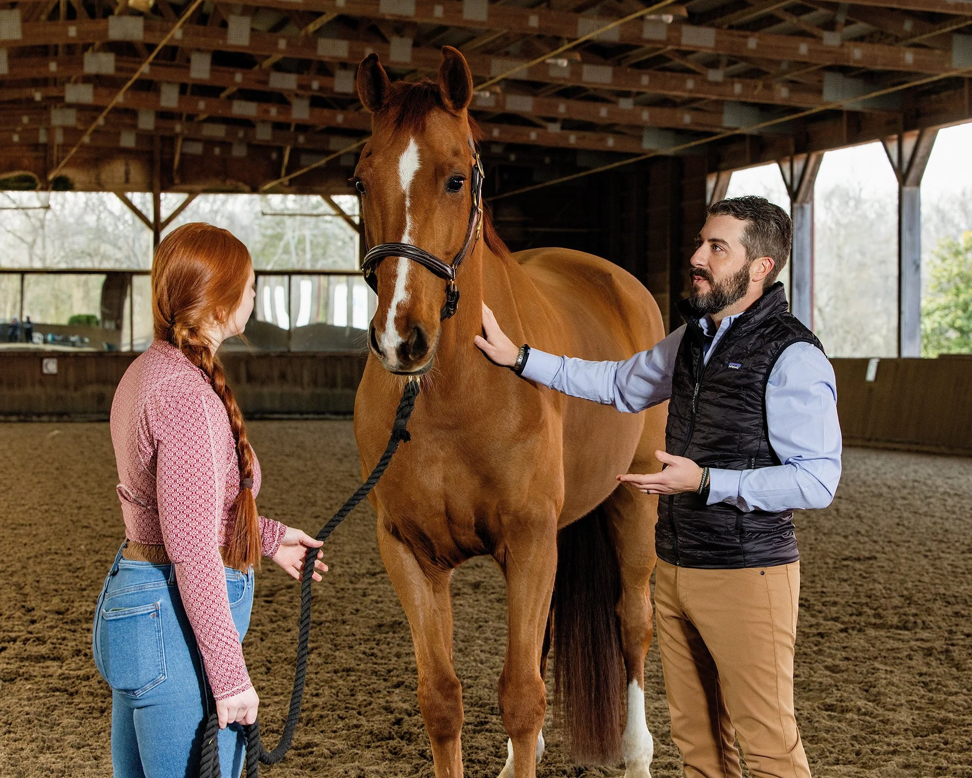 A woman with red hair in a braid, holding a leash attached to a chestnut horse, while a male veterinarian with a beard, dressed in a black vest, gently talks to the woman inside an indoor riding arena with wooden beams.