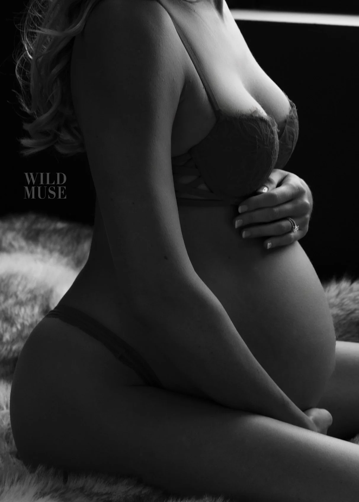 A black and white boudoir photo of a pregnant woman sitting on a furry rug, wearing lingerie, with her hand gently resting on her belly.