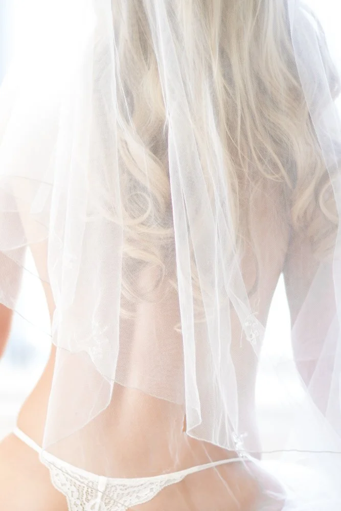 A woman with long blonde hair wearing a sheer white robe and white lace underwear, sitting in a bright, softly lit room.
