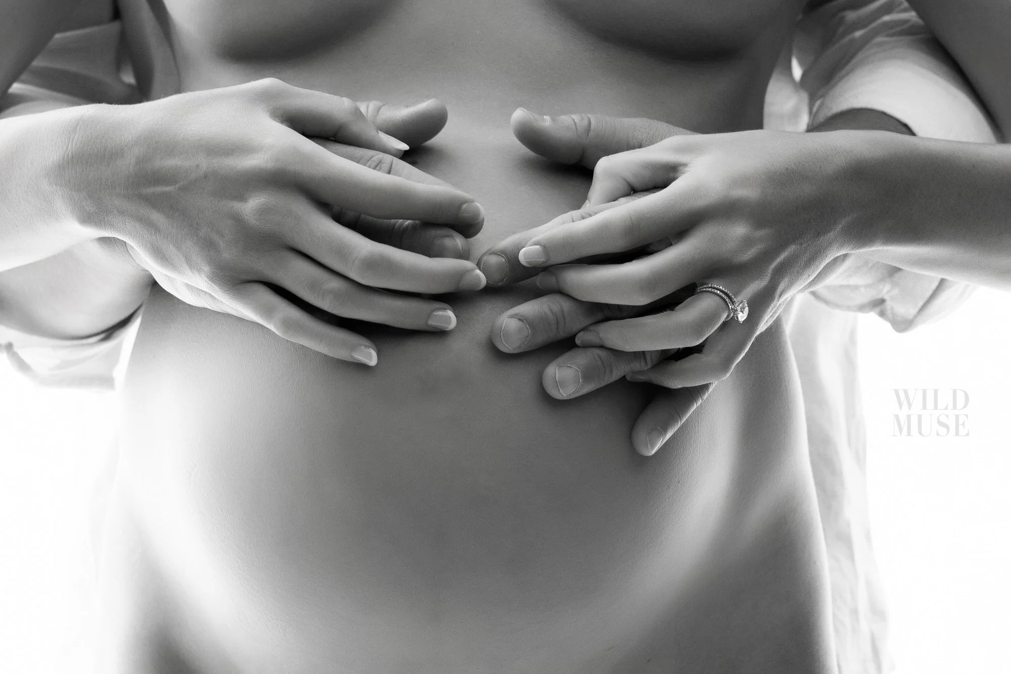 Close-up of two women's hands resting on a pregnant belly, one hand with an engagement ring, in black and white.