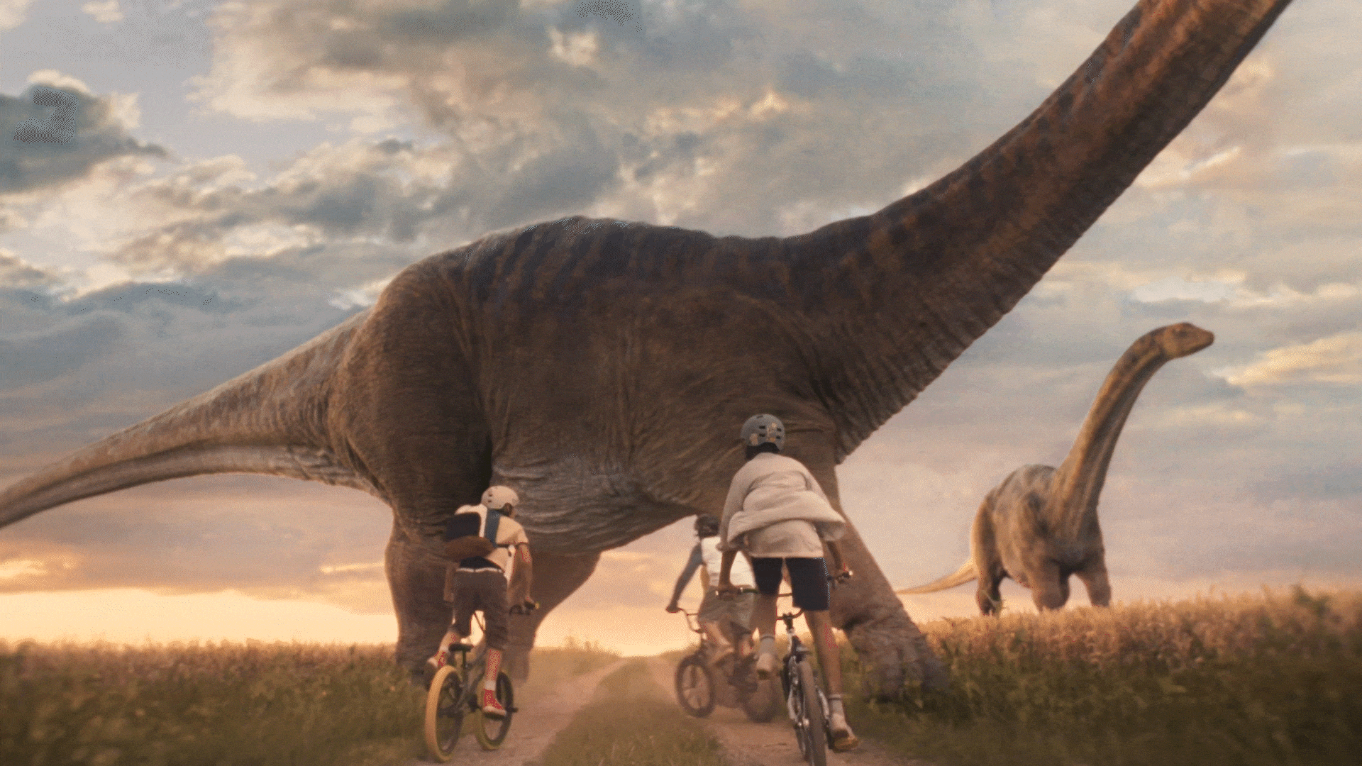 Children riding bikes on a dirt path during sunset, with two large dinosaurs in the background on an open grassy plain.