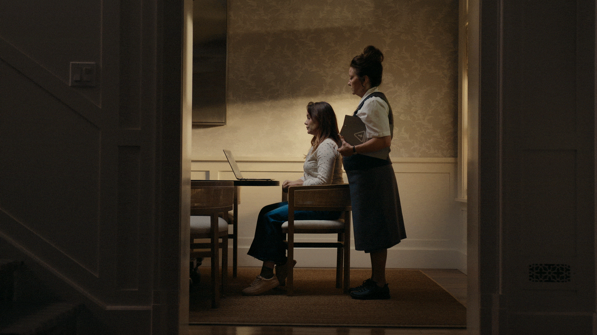 A woman sitting at a dining table using a laptop, with another woman standing beside her holding a notebook and a folder in a dimly lit room.