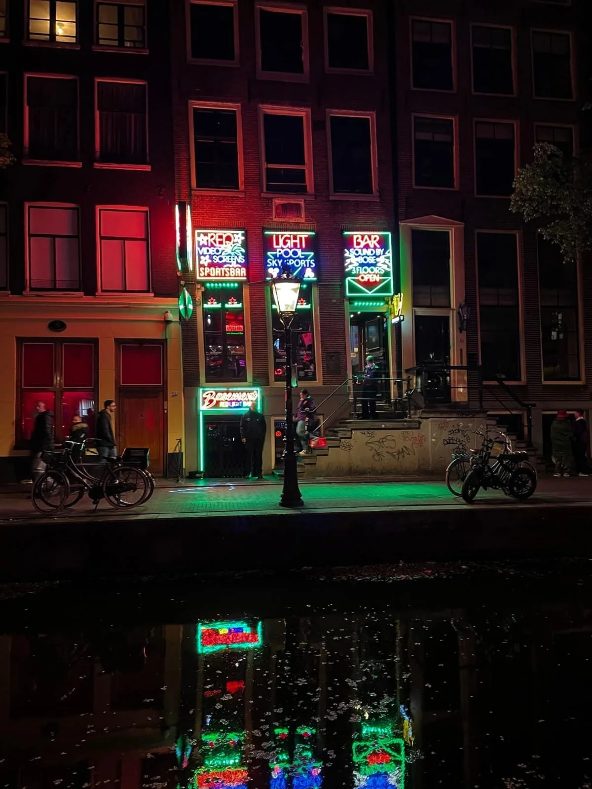 Neon-lit bar in Amsterdam’s Red Light District at night with glowing signage reflected across the canal.