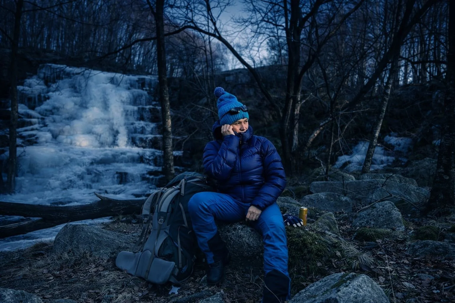 Woman in blue winter clothing sitting on rocks near the frozen waterfall at Sviland Falls in Rogaland, Norway with a hiking backpack during a February outdoor trek.